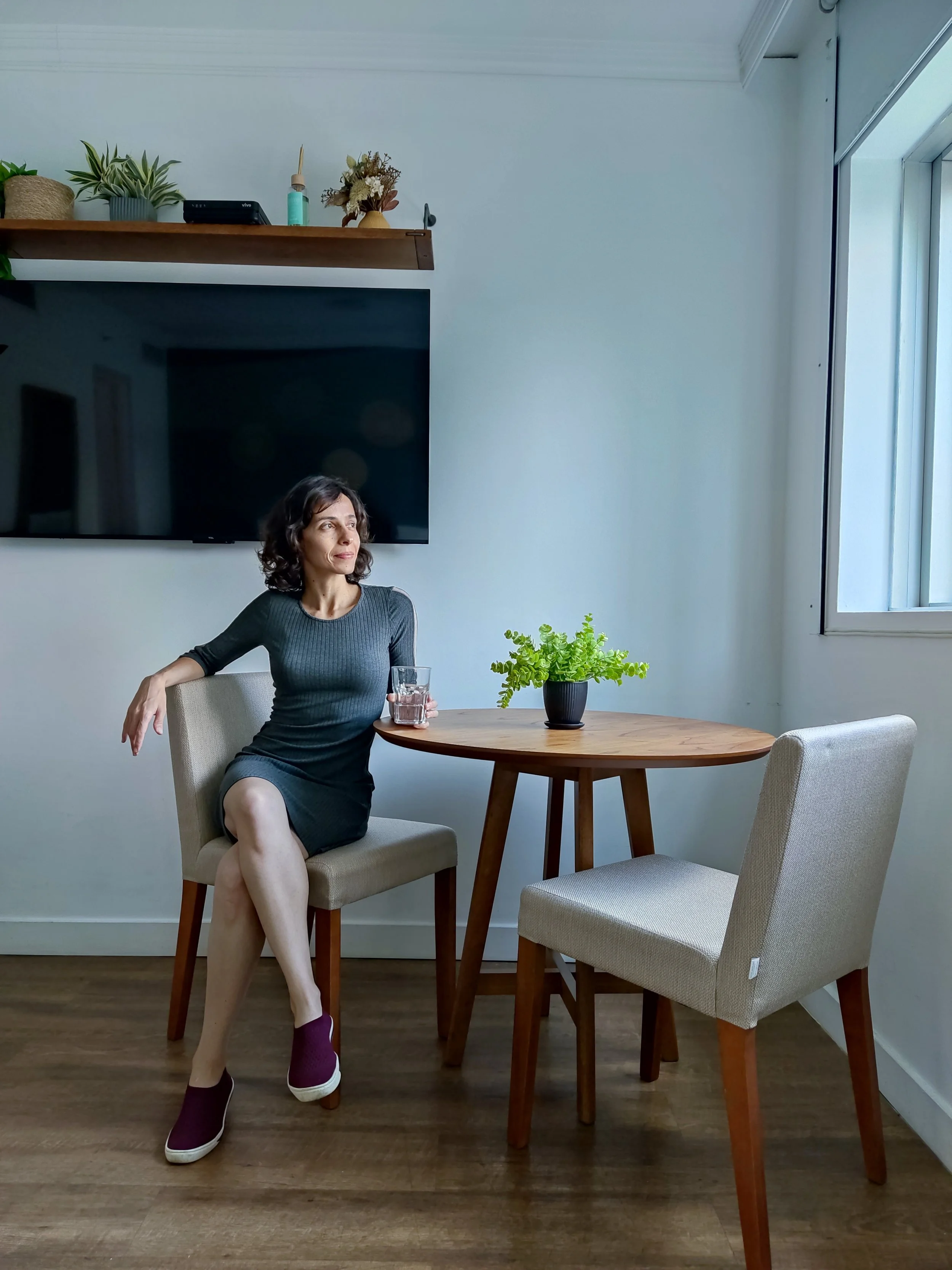 Mulher sentada com uma taça de água na mesa, no ambiente de uma sala com televisão, plantas e janela, decorada de forma moderna e minimalista.