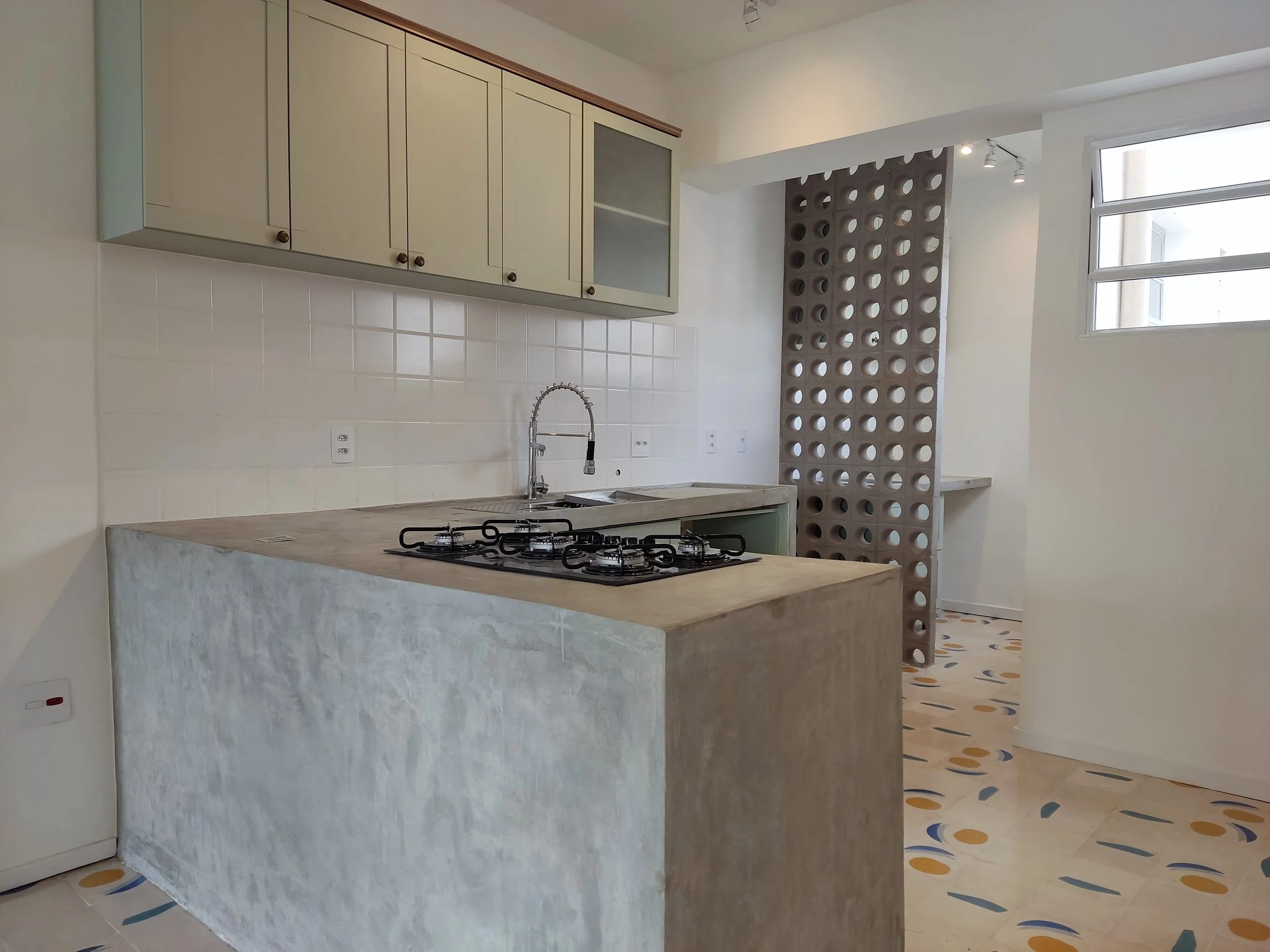 Cozinha moderna com balcão de concreto, pia, pia, armários suspensos em tom claro, parede de azulejos brancos, janela grande, divisão com painel de blocos de concreto com buracos.
