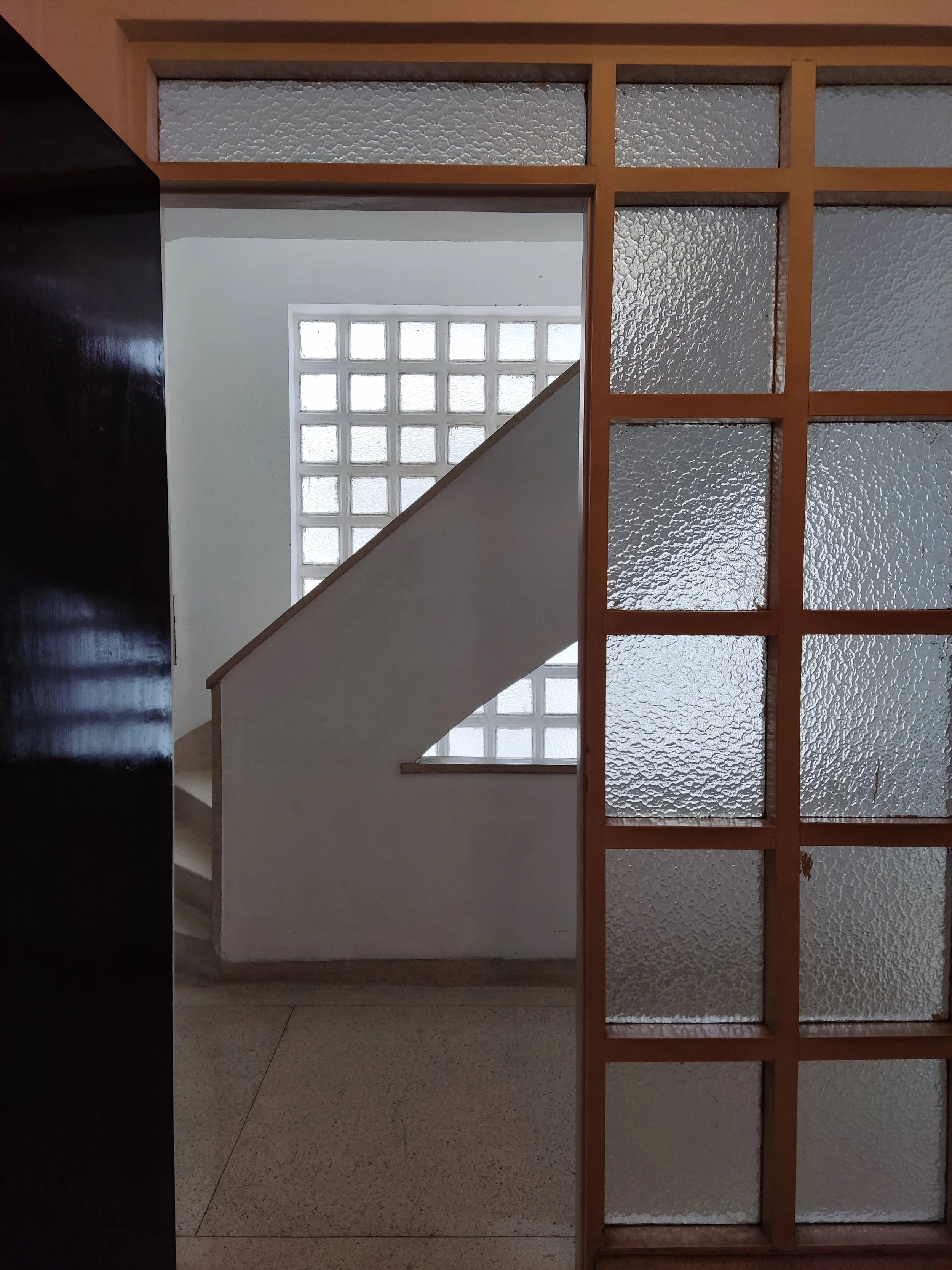 Entrada de um edifício, com porta de vidro fosco, mostrando uma escada branca ao fundo e uma janela de blocos de vidro na parede ao lado.