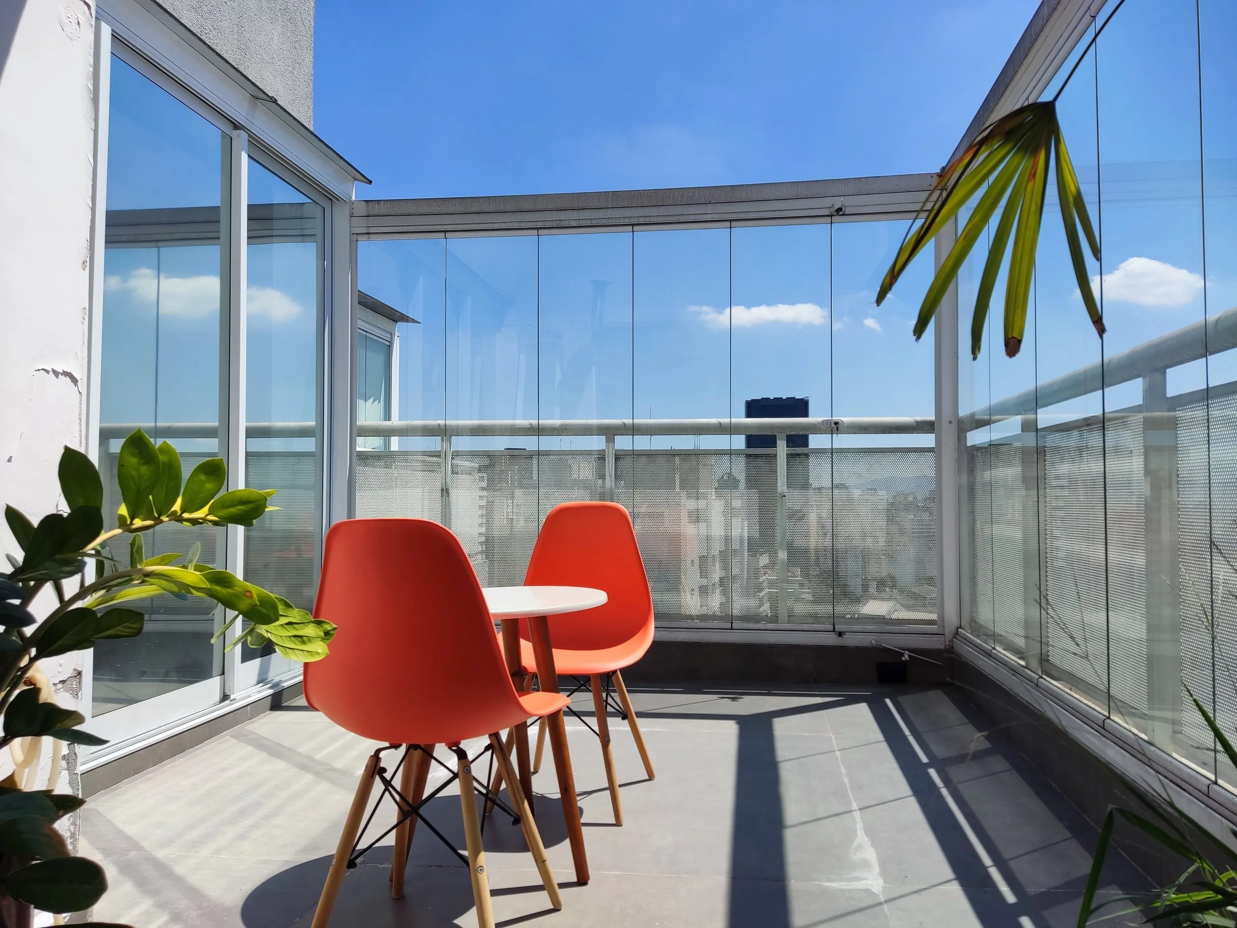 Varanda com duas cadeiras laranja e uma mesa branca, cercada por vidro, com plantas verdes ao redor e vista para o céu azul com algumas nuvens.