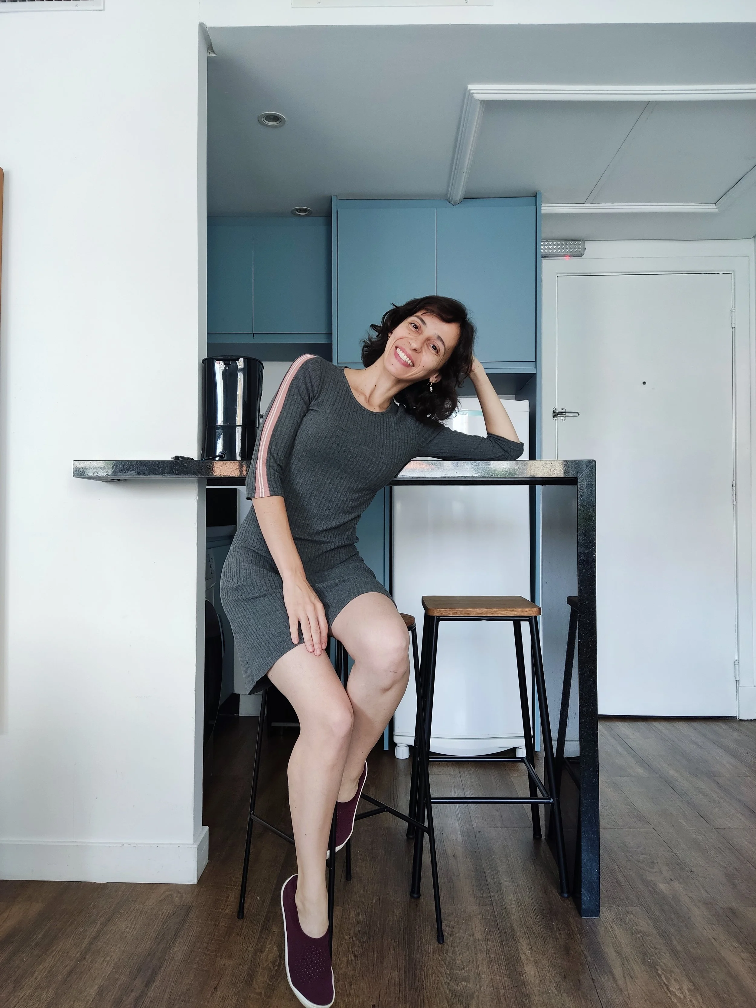 Mulher sorridente sentada numa bancada de cozinha, descansando a cabeça na mão, usando vestido cinza escuro e tênis vinho, com fundo de armários azuis e piso de madeira.