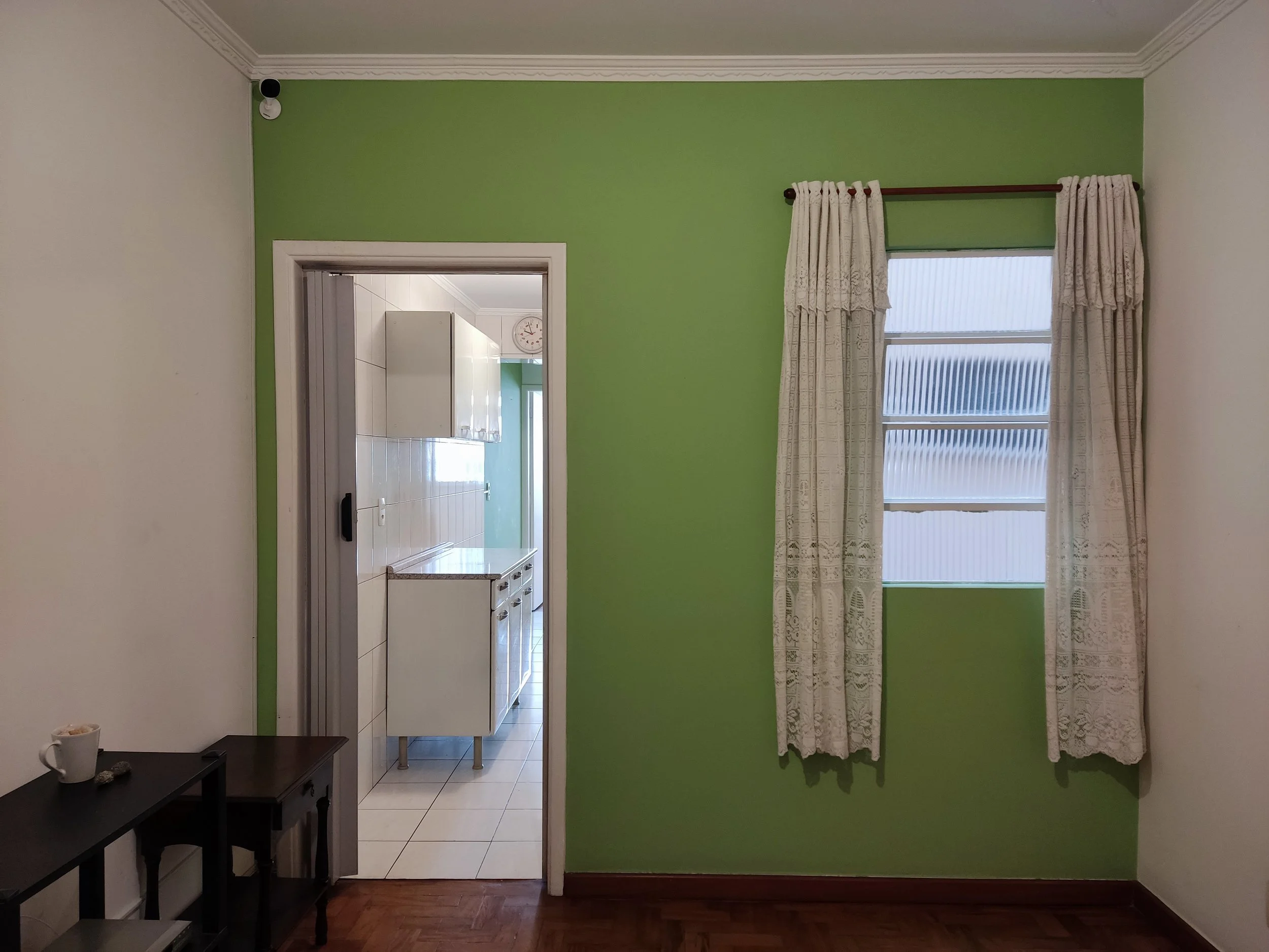Sala com parede verde, janela com cortinas brancas e cozinha visível ao fundo, com piso de azulejos brancos e armários brancos.