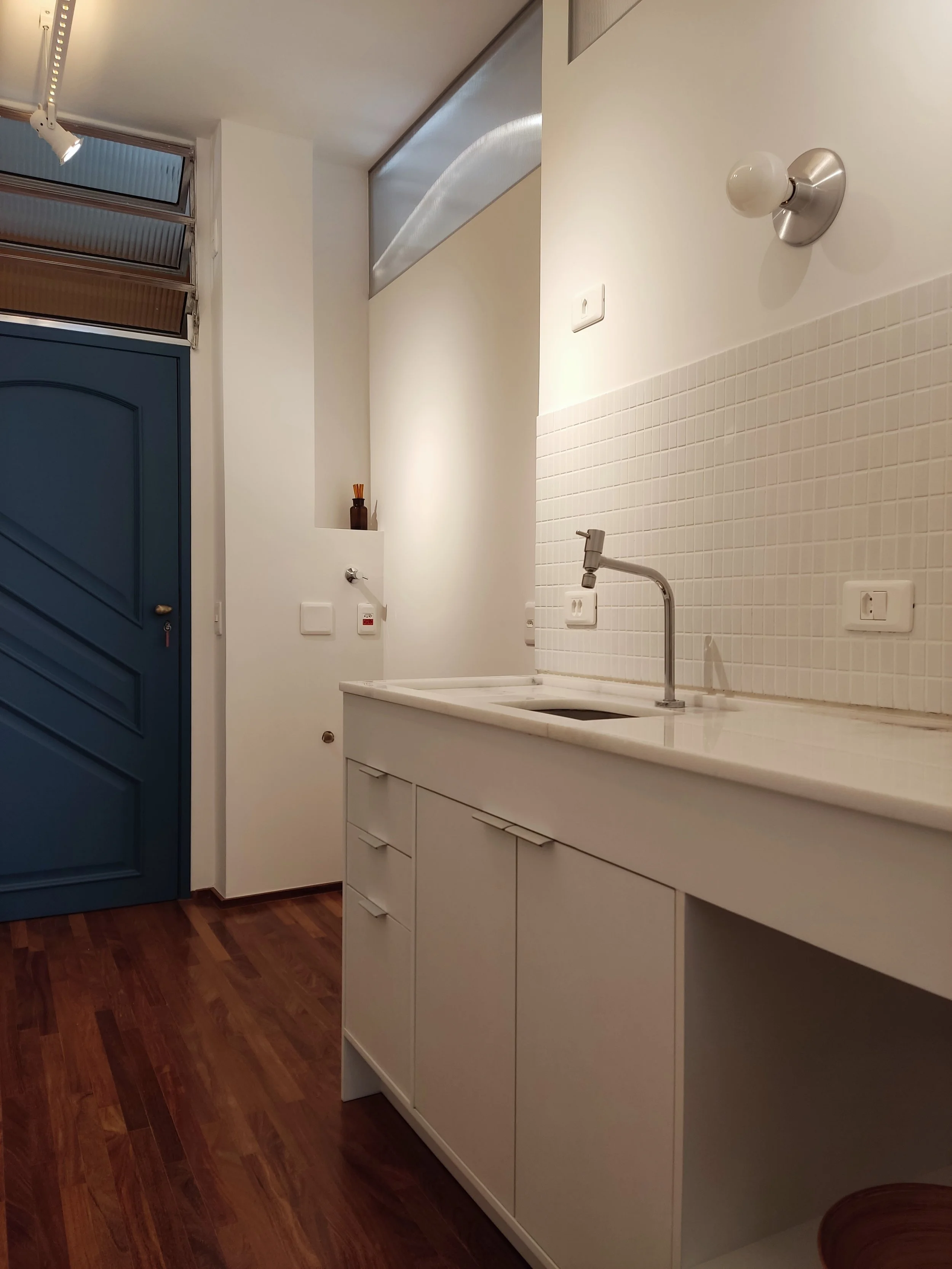 Cozinha com bancada branca, azulejos brancos, uma torneira de pia e uma parede branca com uma luz de teto