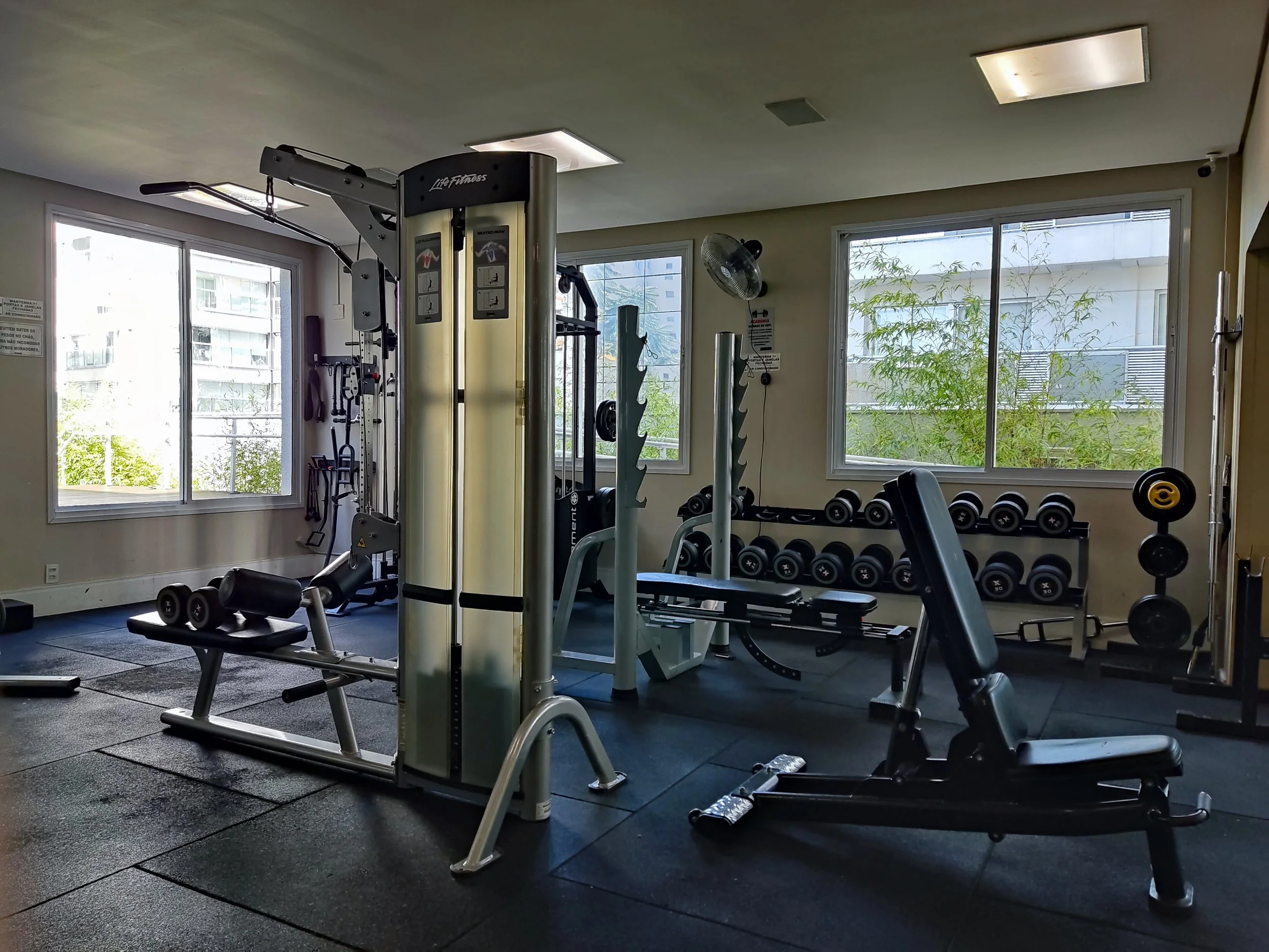 Sala de ginástica com equipamentos de musculação, pesos livres, e máquinas de treino, tendo janelas que deixam entrar luz natural.