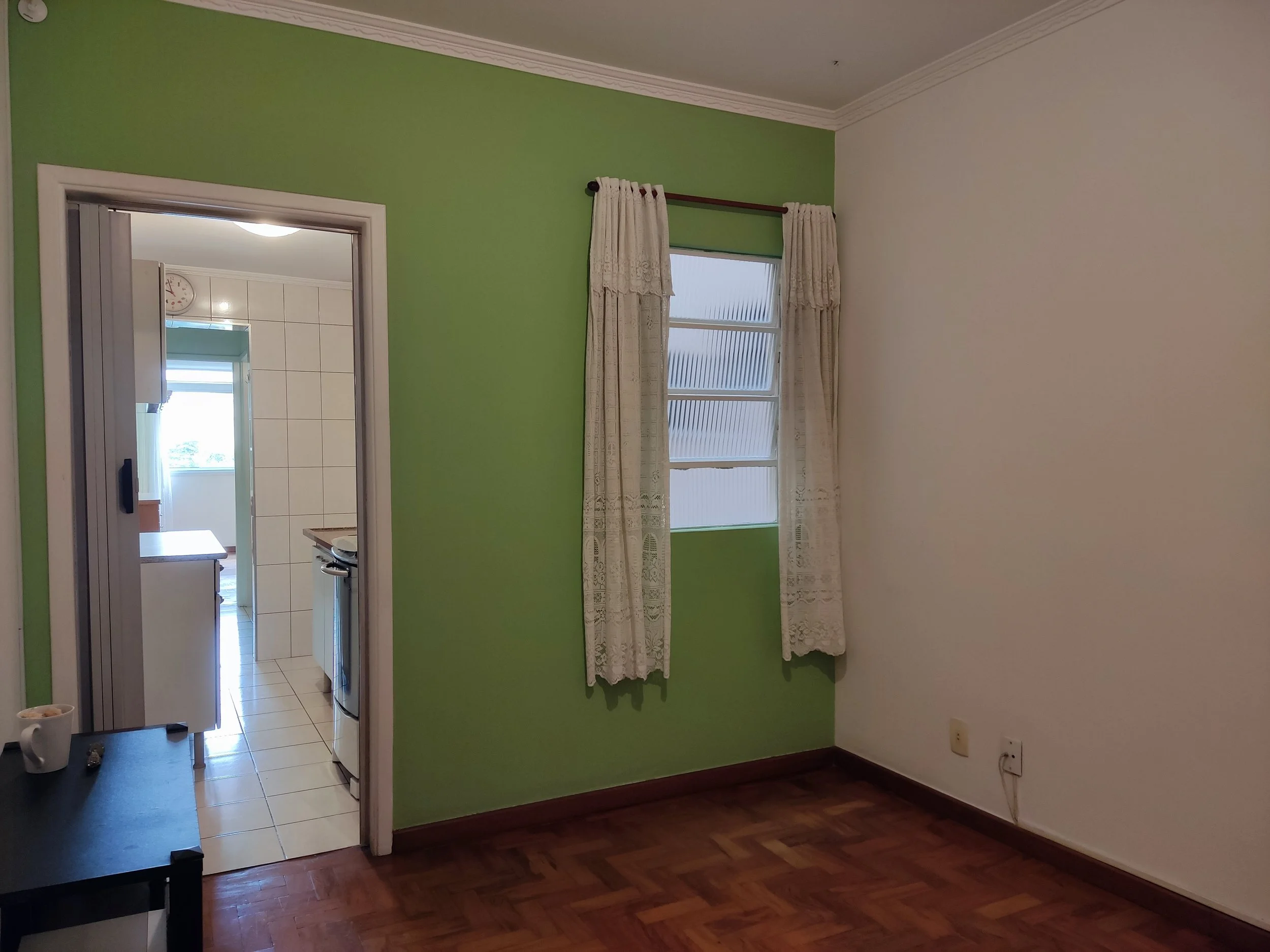 Quarto com parede branca e parede verde, janela com cortinas bege, piso de madeira escura, porta de entrada para a cozinha com azulejos brancos e eletrodomésticos visíveis.