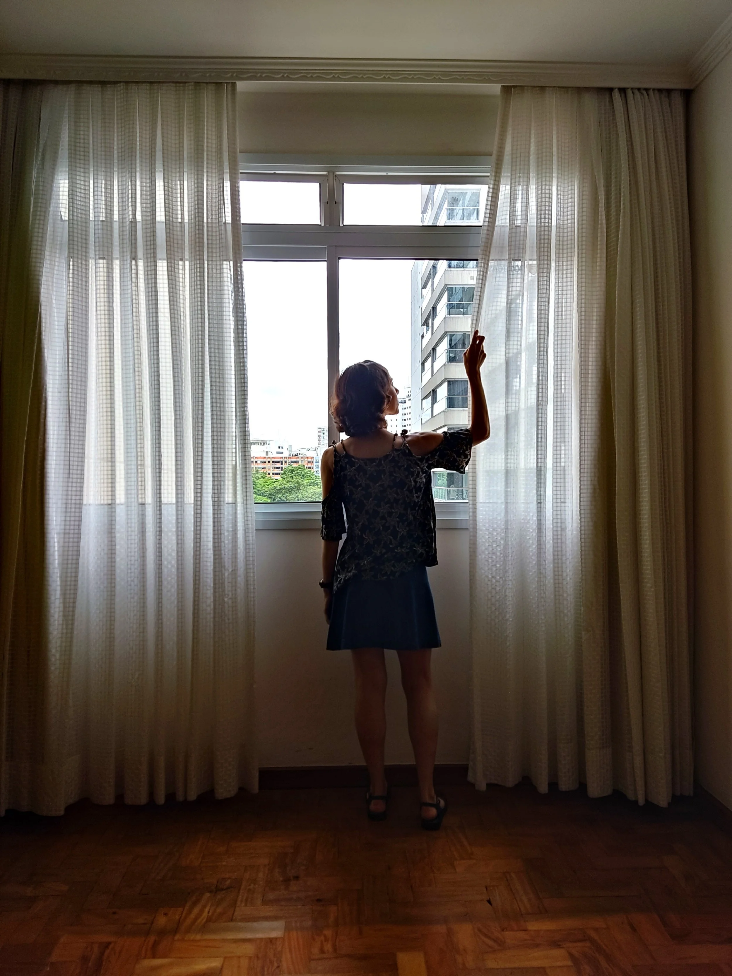 Mulher de costas, de vestido preto com estampa, de cabelo curto, ajustando as cortinas brancas em uma sala com janelas amplas, chão de madeira e prédio de edifício alto ao fundo.