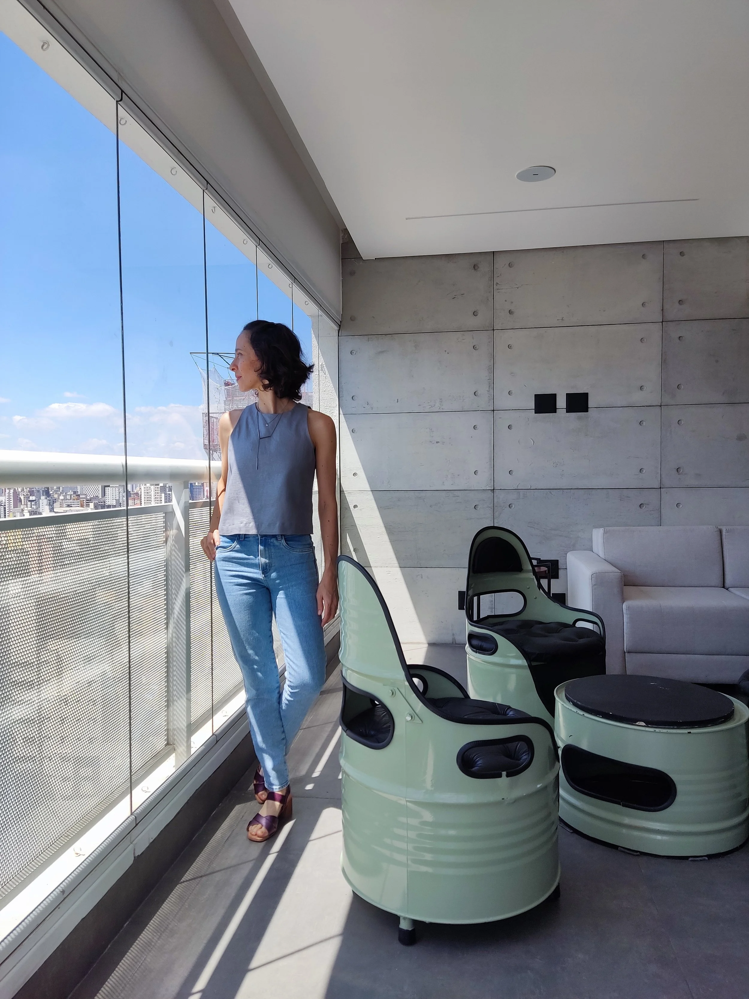 Mulher de cabelos escuros e roupa cinza, de pé ao lado de uma varanda de um prédio, olhando para fora em um dia ensolarado, com vista para a cidade.