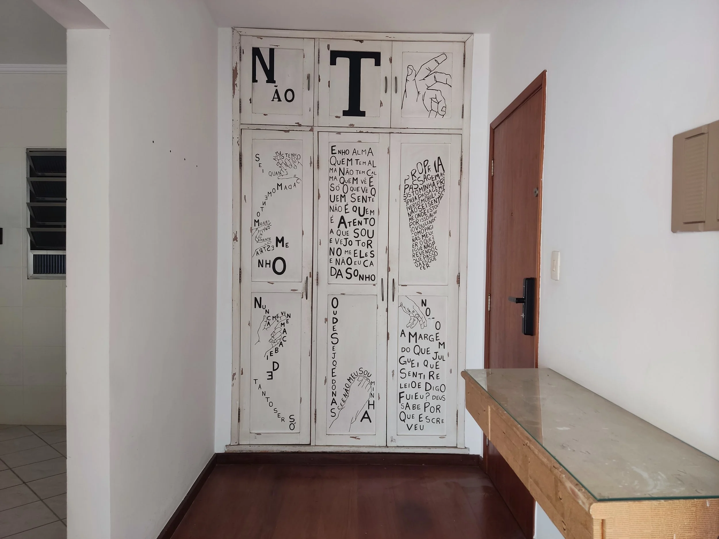 Armário de madeira branco com detalhes de letras e desenhos feitos à mão, com várias frases e palavras em português, localizado em uma parede branca. Ao lado, uma porta de madeira escura e uma mesa com tampo de vidro na parede direita.