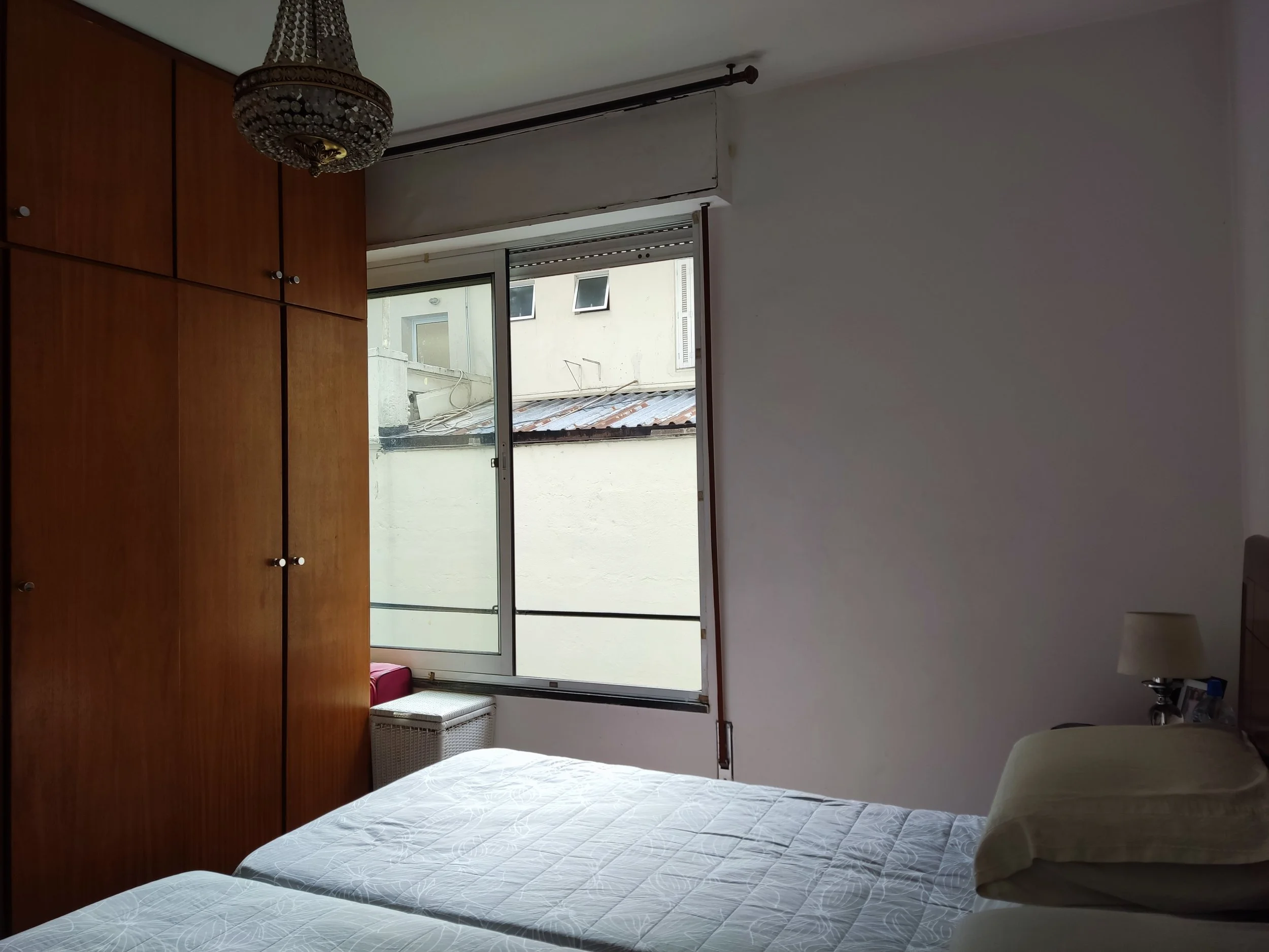 Quarto com duas camas de casal, armário de madeira grande, janela aberta mostrando prédio vizinho, cômoda com luminária ao lado da cama.