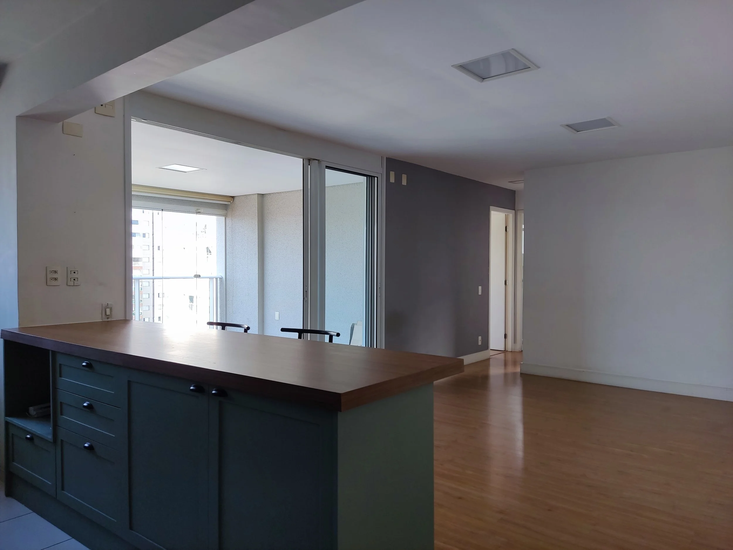 Sala de estar vazia com chão de madeira, parede branca e parede cinza escura, com uma porta aberta, uma janela de vidro com varanda e iluminação do teto.