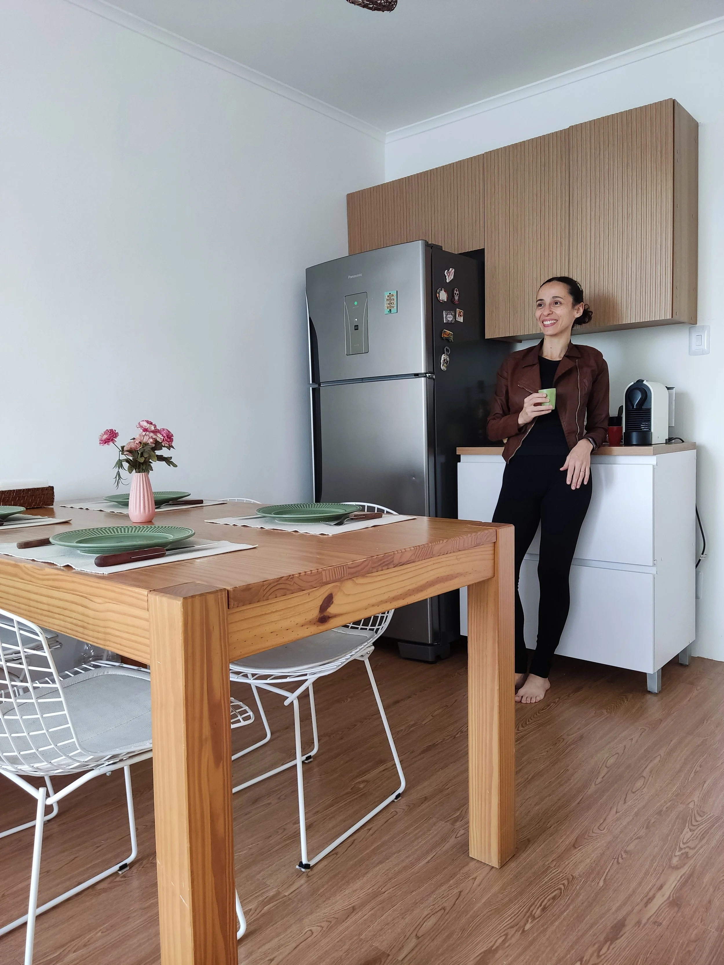 Mulher sorridente com blusa preta e jaqueta marrom, segurando uma caneca, ao lado de uma cozinha com geladeira de inox, armários de madeira e mesa de jantar de madeira com pratos verdes e guardanapos marrons.