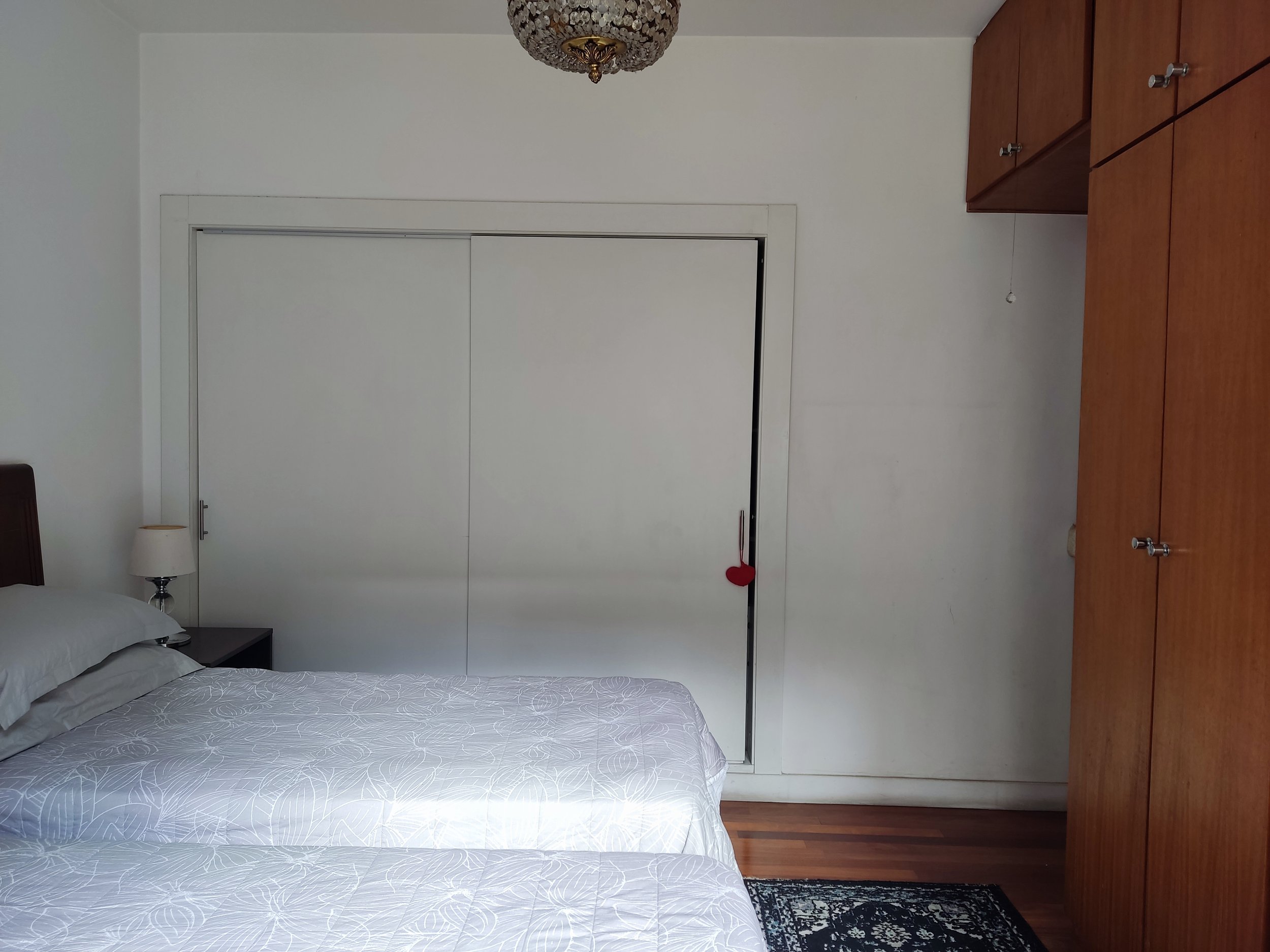 quarto com duas camas de solteiro, criado-mudo com abajur, guarda-roupa de madeira, porta de correr branca com coração decorativo, piso de madeira, teto com luminária decorativa.