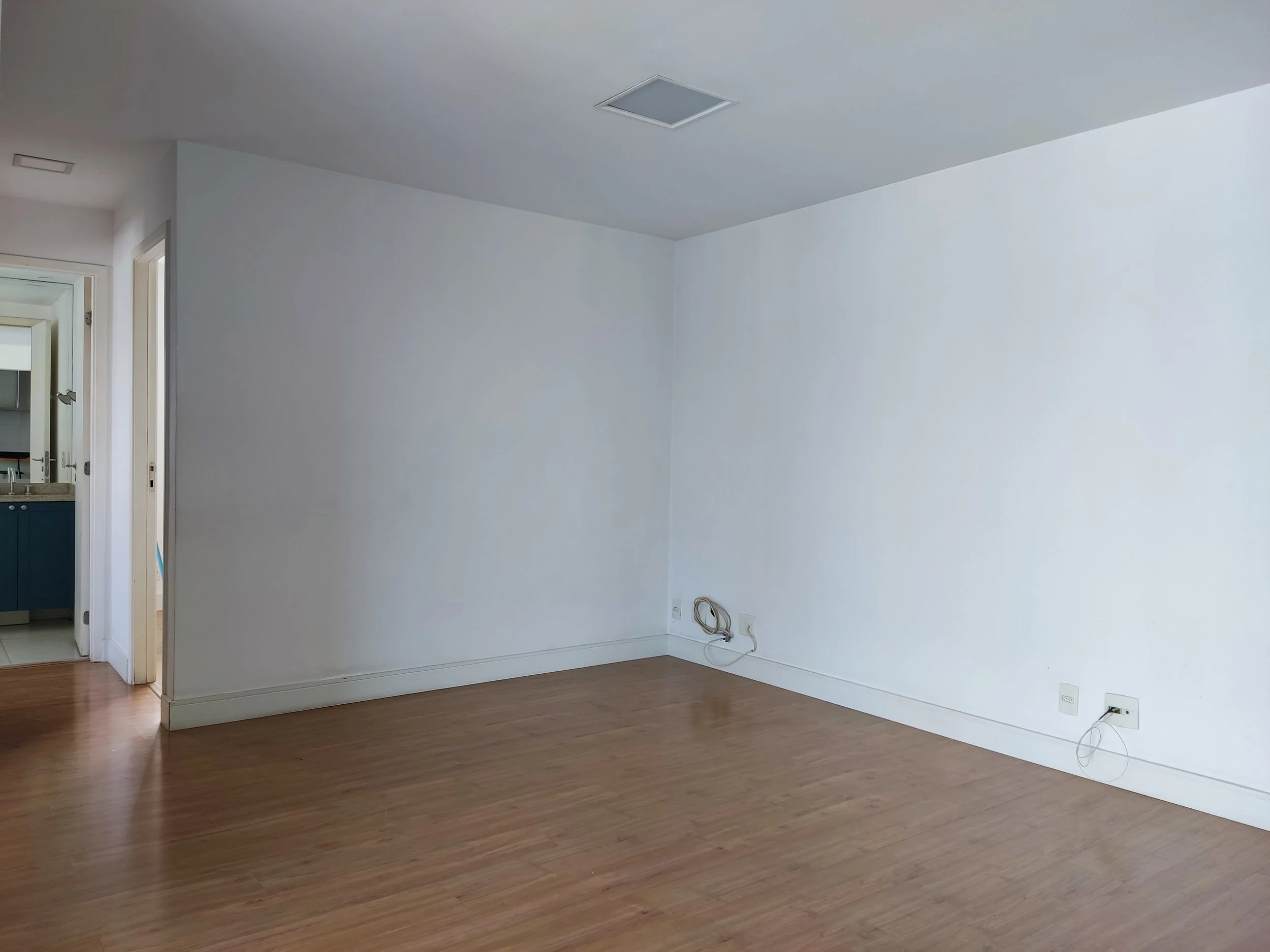 Sala vazia com piso de madeira, paredes brancas e uma porta que leva à cozinha, tomadas na parede com cabos de energia.