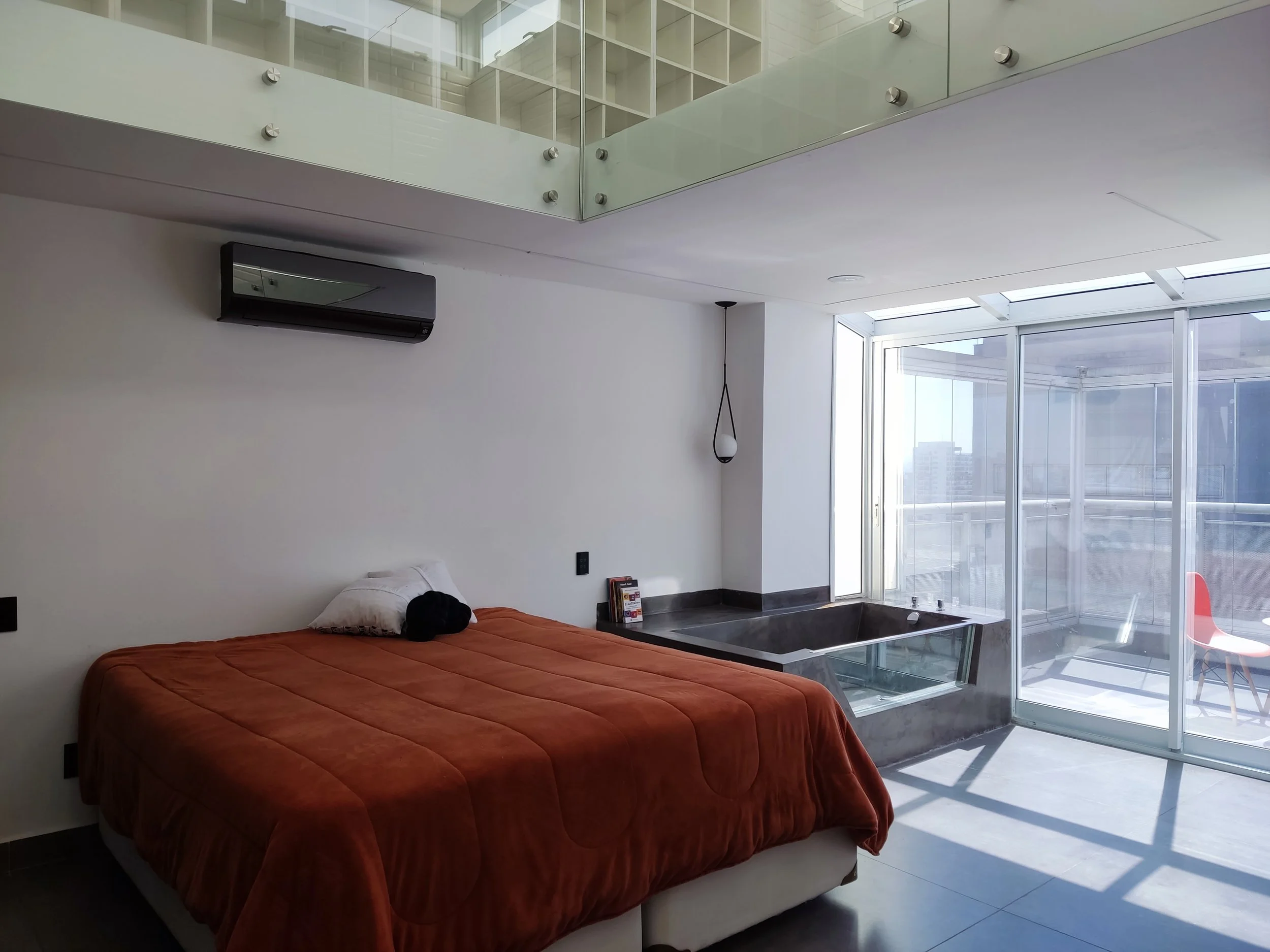 Quarto moderno com cama de roupa de cama laranja, janela grande com varanda, banheira embutida, ar-condicionado na parede, decoração minimalista, lampada pendente e vista da cidade.