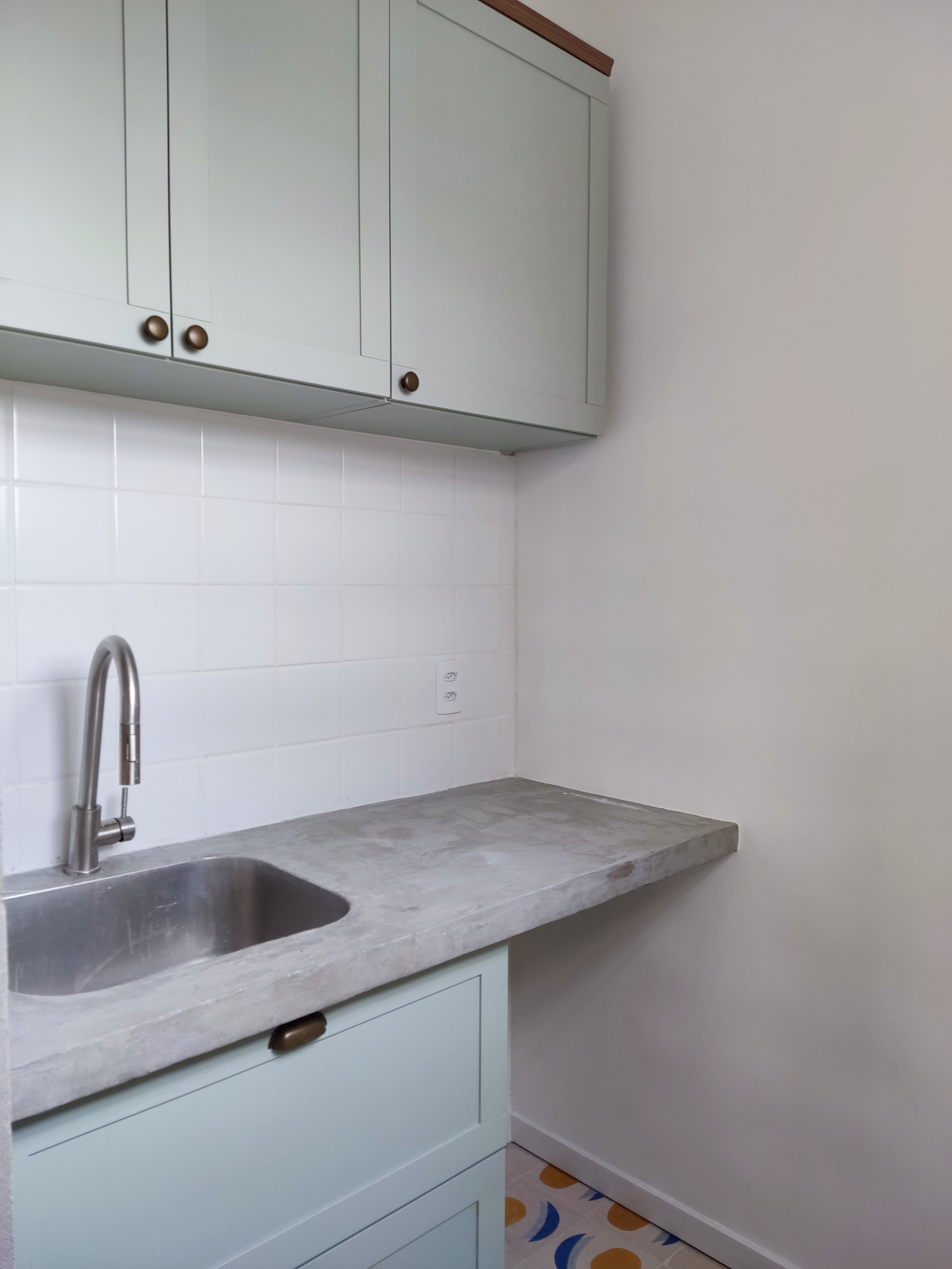 Cozinha com pia de pia de aço inox e bancada de concreto, armário fino de gaveta e armário superior em tom de cinza claro, com piso com azulejos decorativos coloridos.