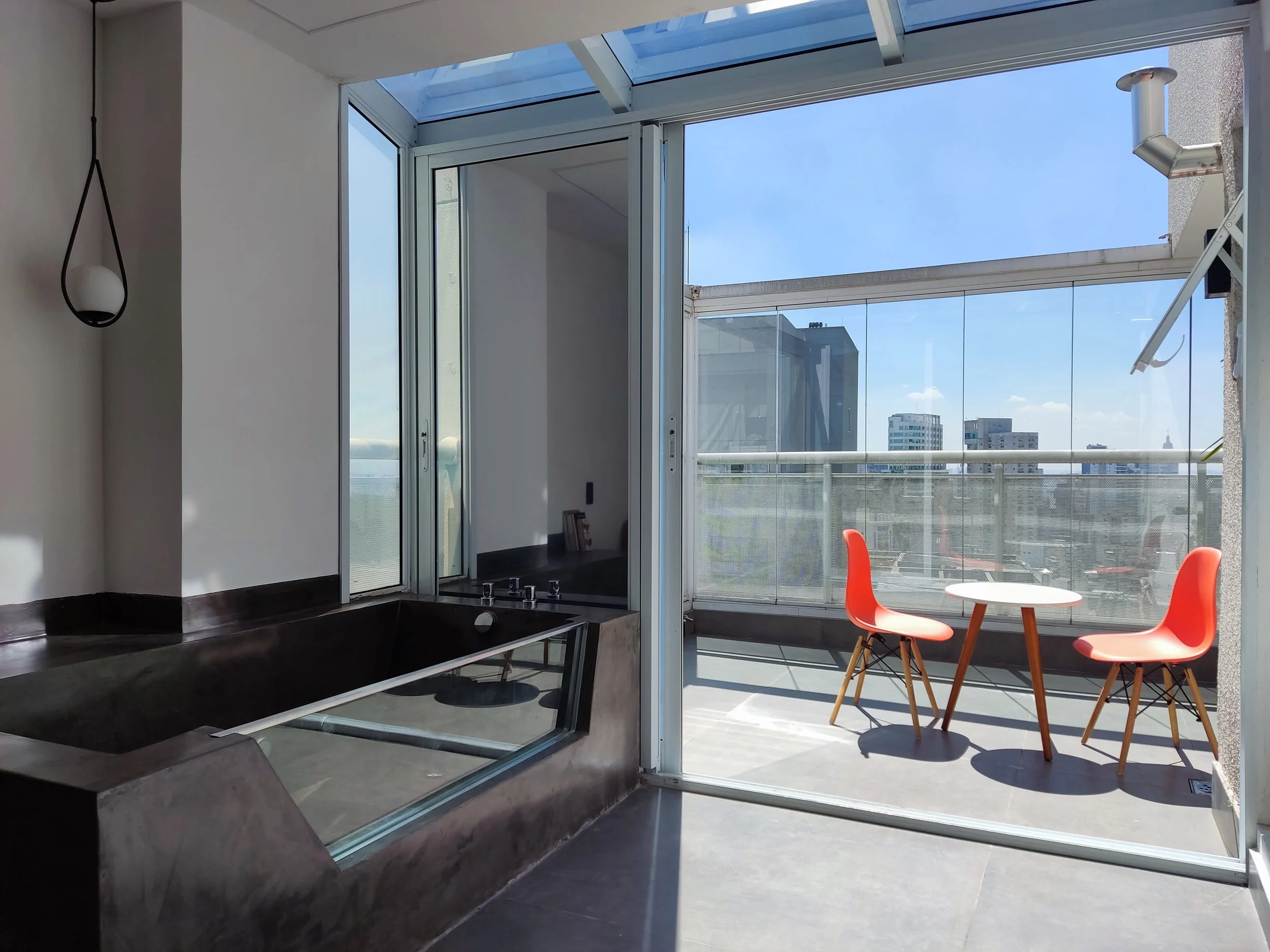 Espaço com banheira de hidromassagem ao lado de uma porta de vidro que leva a uma varanda com duas cadeiras vermelhas e uma mesa redonda sob um teto de vidro, com vista para prédios e céu azul.