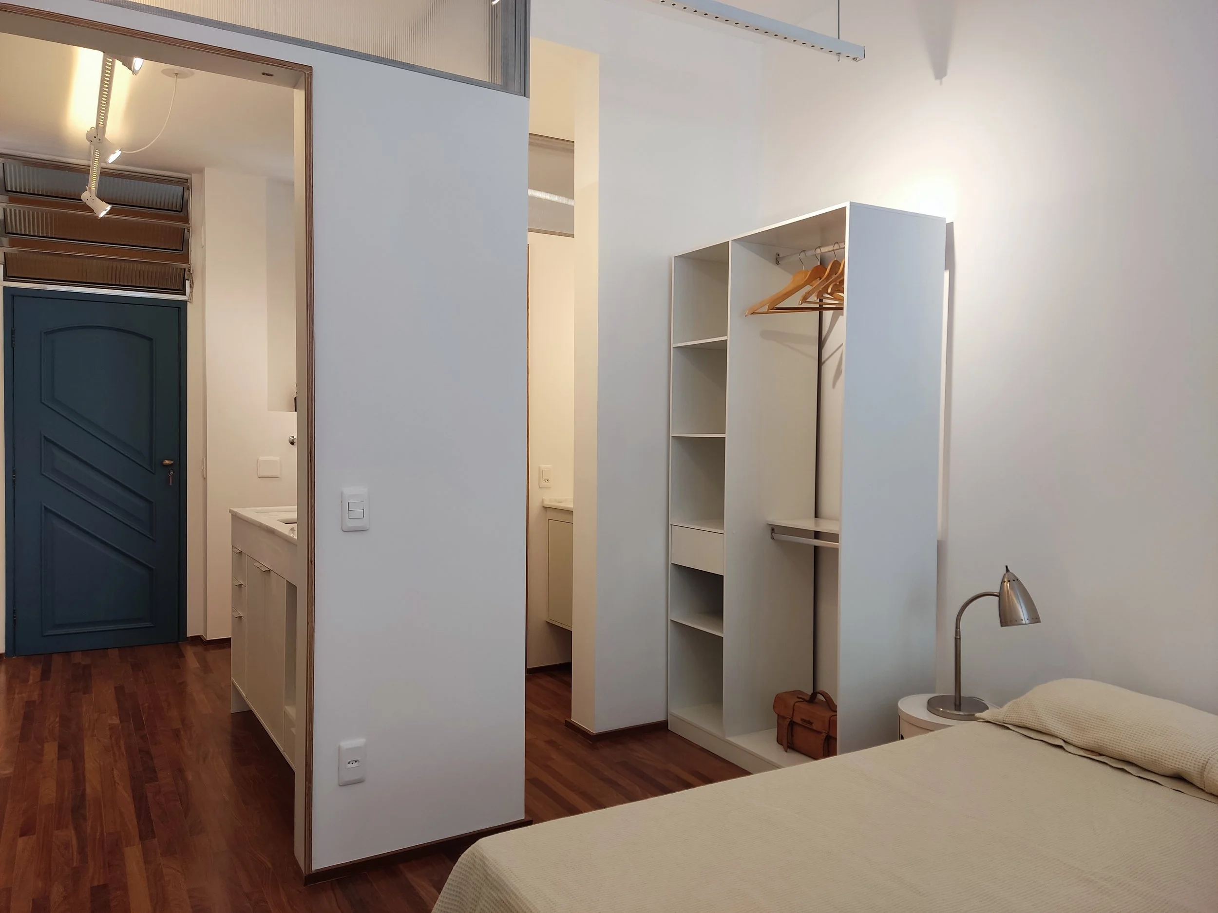 Quarto com cama de roupa de cama bege, luminária de mesa ao lado da cama, armário branco aberto com cabides de roupas e uma pequena bolsa, piso de madeira escura, parede branca, porta azul na entrada, teto com iluminação embutida.