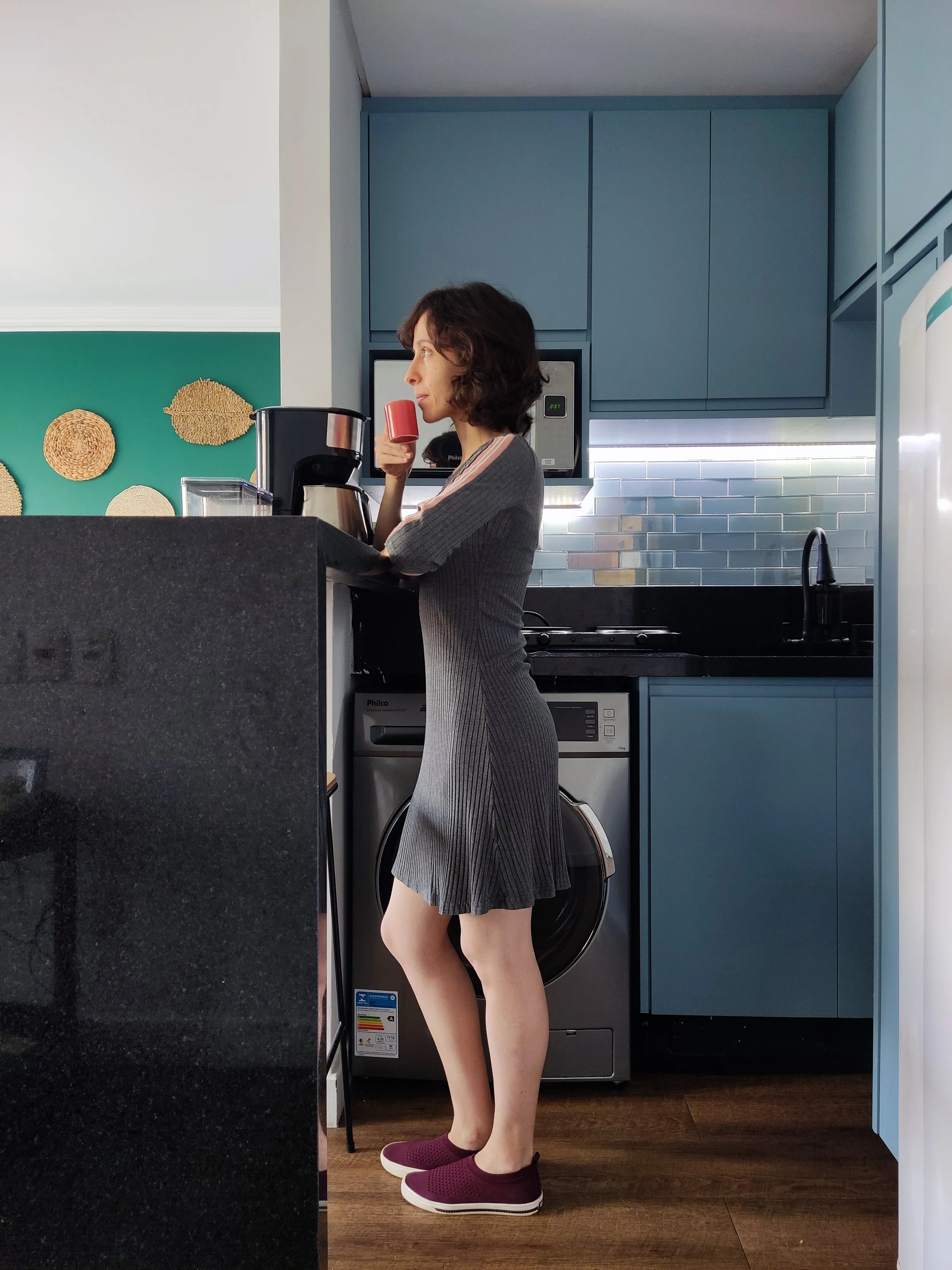 Mulher com cabelo cacheado, vestindo vestido cinza de manga longa, de roupa descalça, tomando café na cozinha moderna com armários azuis e paredes com revestimento de azulejos. Ela está de perfil, ao lado de uma máquina de lavar e ao lado de um balcã