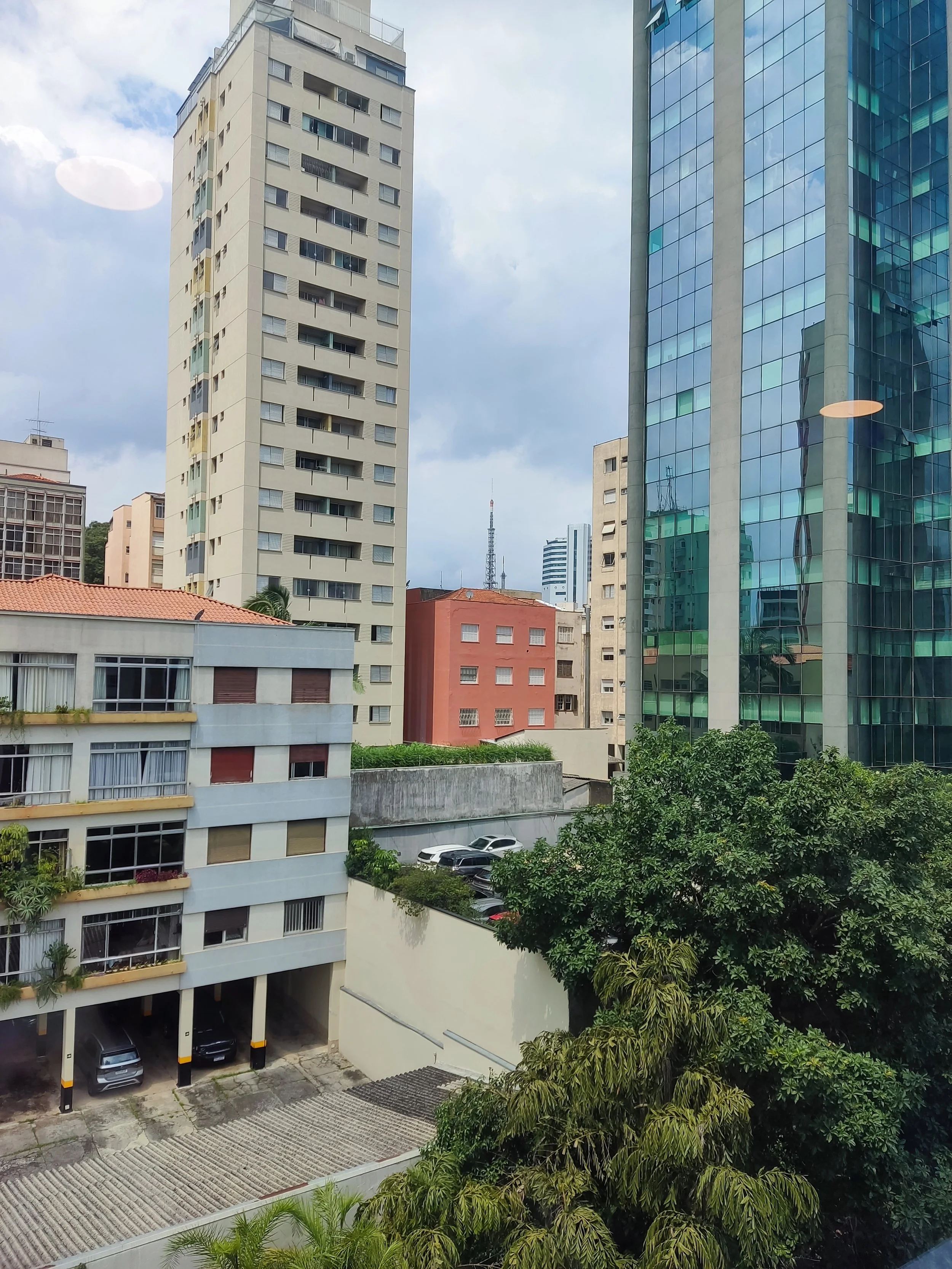 Cena de uma cidade com prédios altos, incluindo um prédio branco de janelas pequenas e um edifício de vidro reflexivo. Há árvores verdes na parte inferior da imagem e carros estacionados ao lado de um estacionamento no canto inferior esquerdo. O céu 