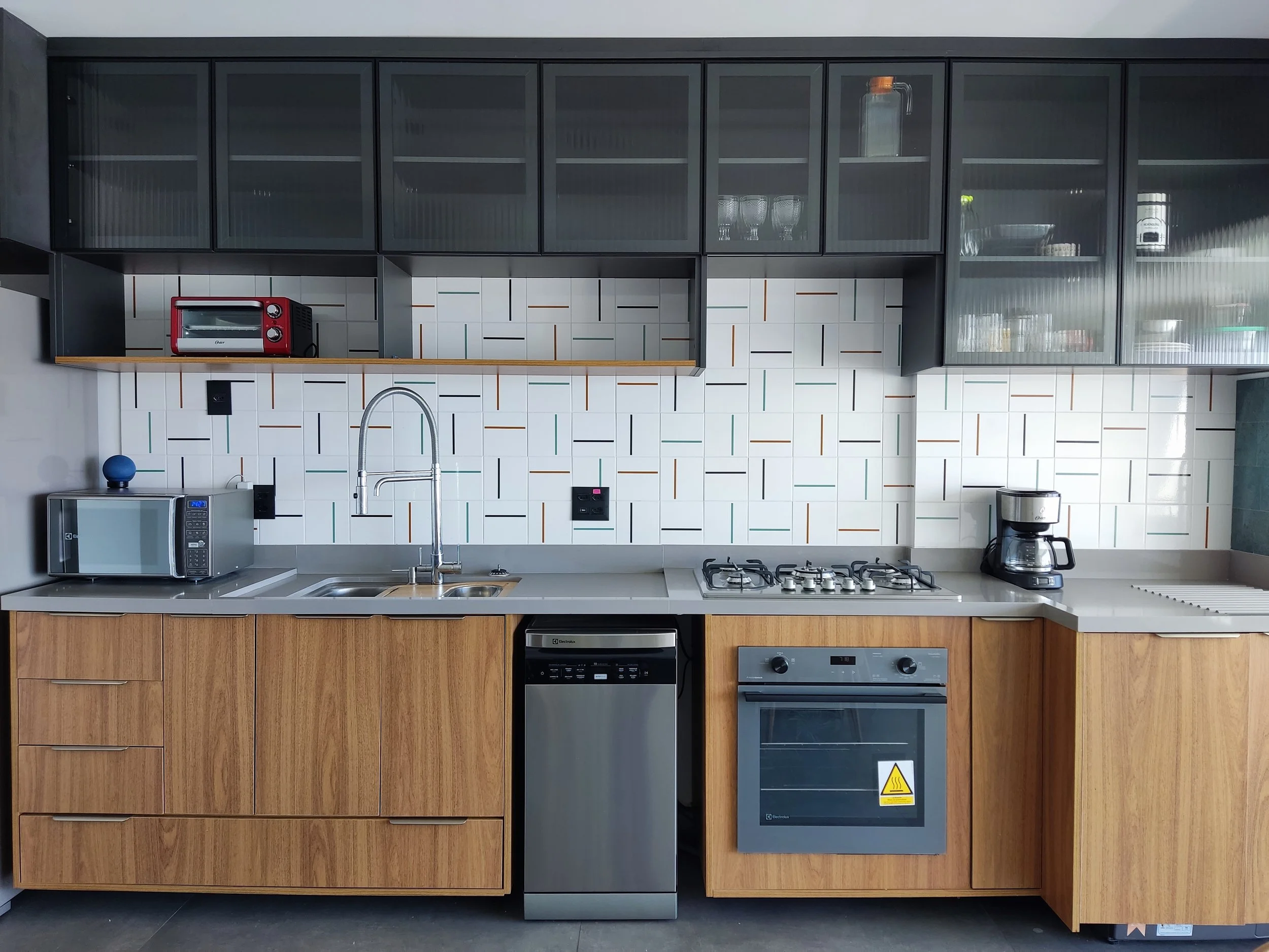 Cozinha moderna com armários de madeira na parte inferior, bancada, pia com torneira, geladeira, forno embutido, micro-ondas, café na máquina, forno de micro-ondas em altura superior, armários de vidro na parte superior e parede decorada com revestim