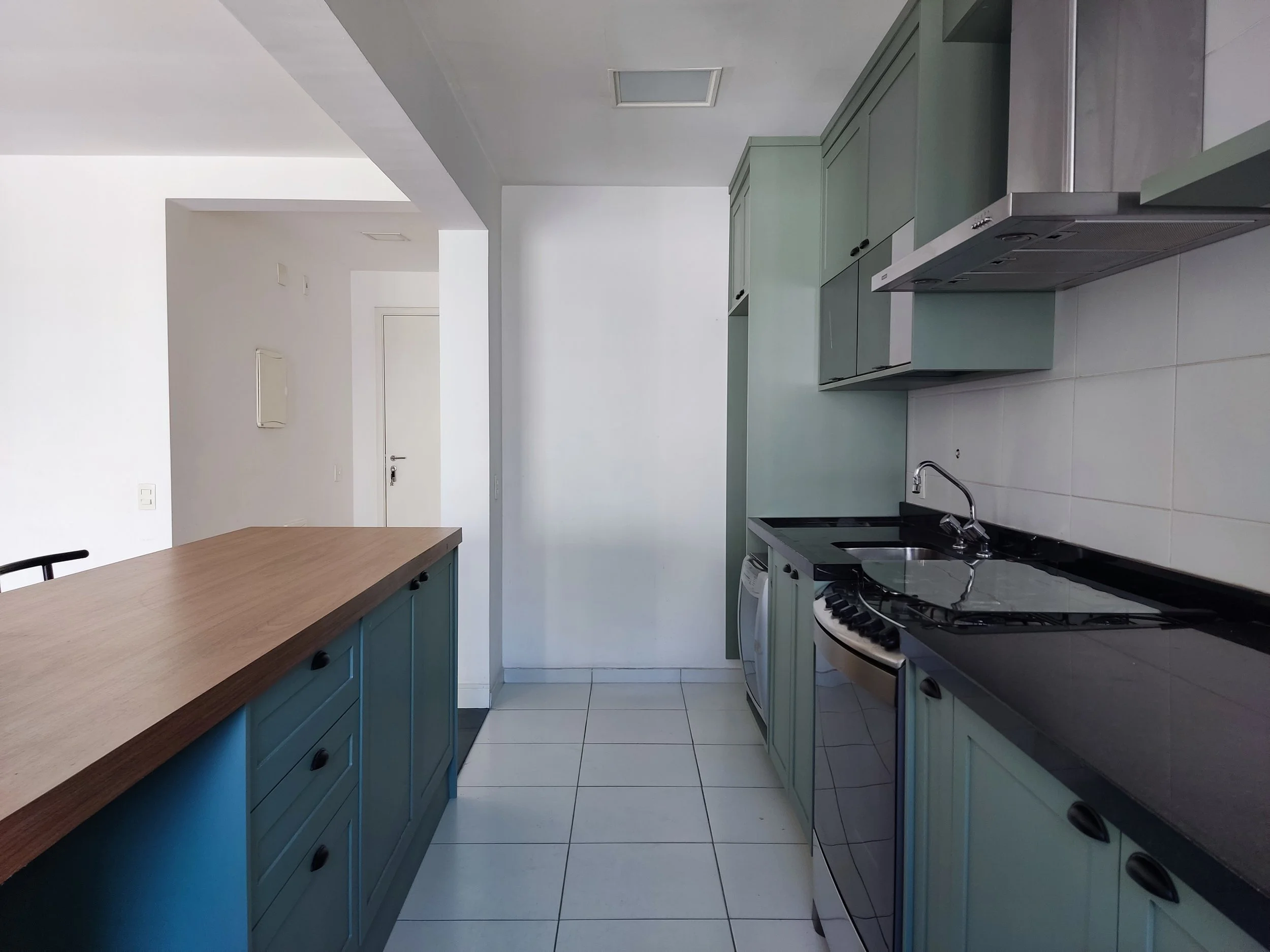 Cozinha moderna com armários verdes, balcão de madeira, pia, fogão e forno, piso de azulejo branco, paredes brancas e iluminação natural.