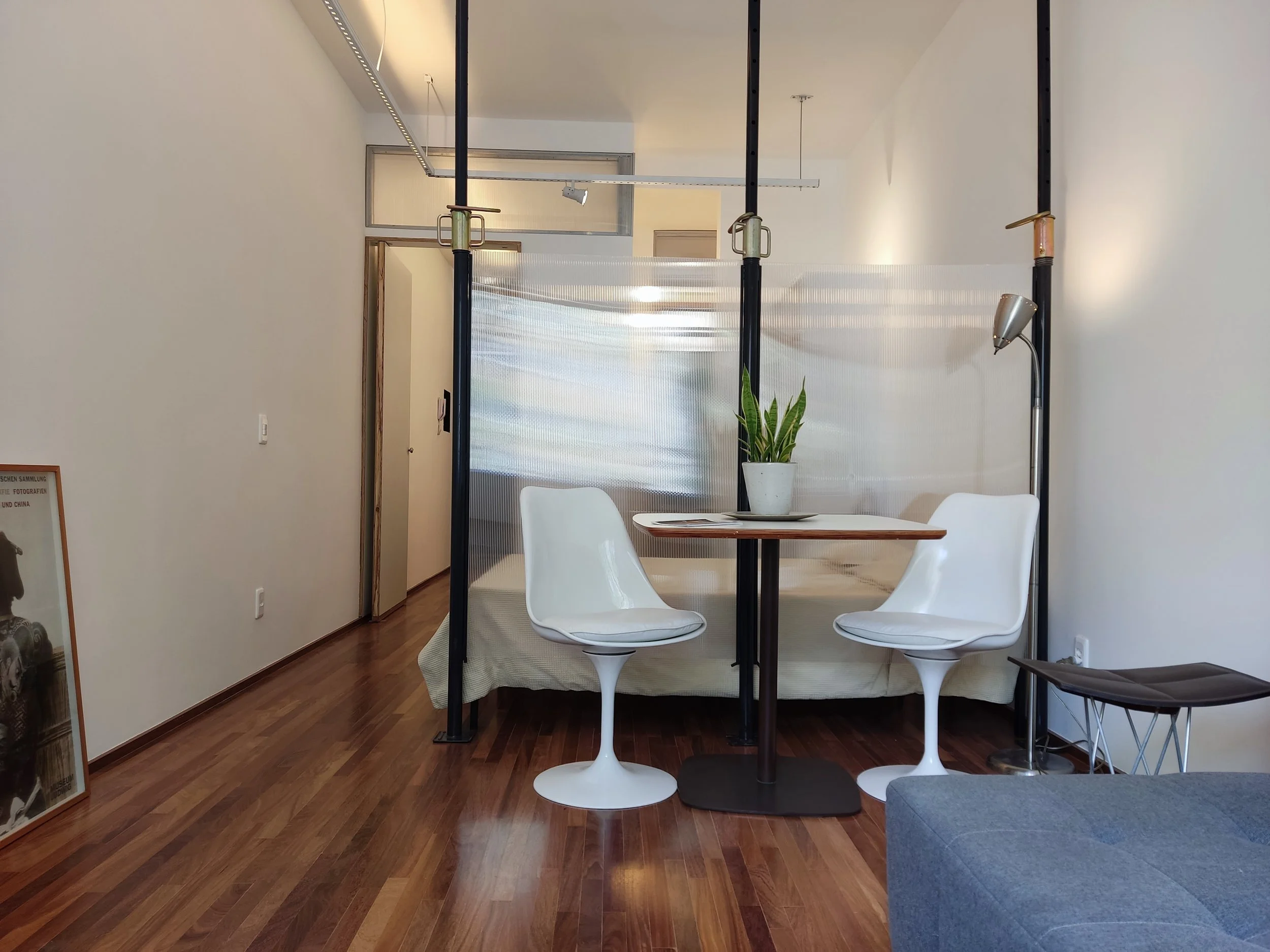 Sala com duas cadeiras brancas ao redor de uma mesa redonda de madeira, uma planta em vaso na mesa, parede branca, piso de madeira, com parte de sofá cinza à direita e uma iluminação de teto.