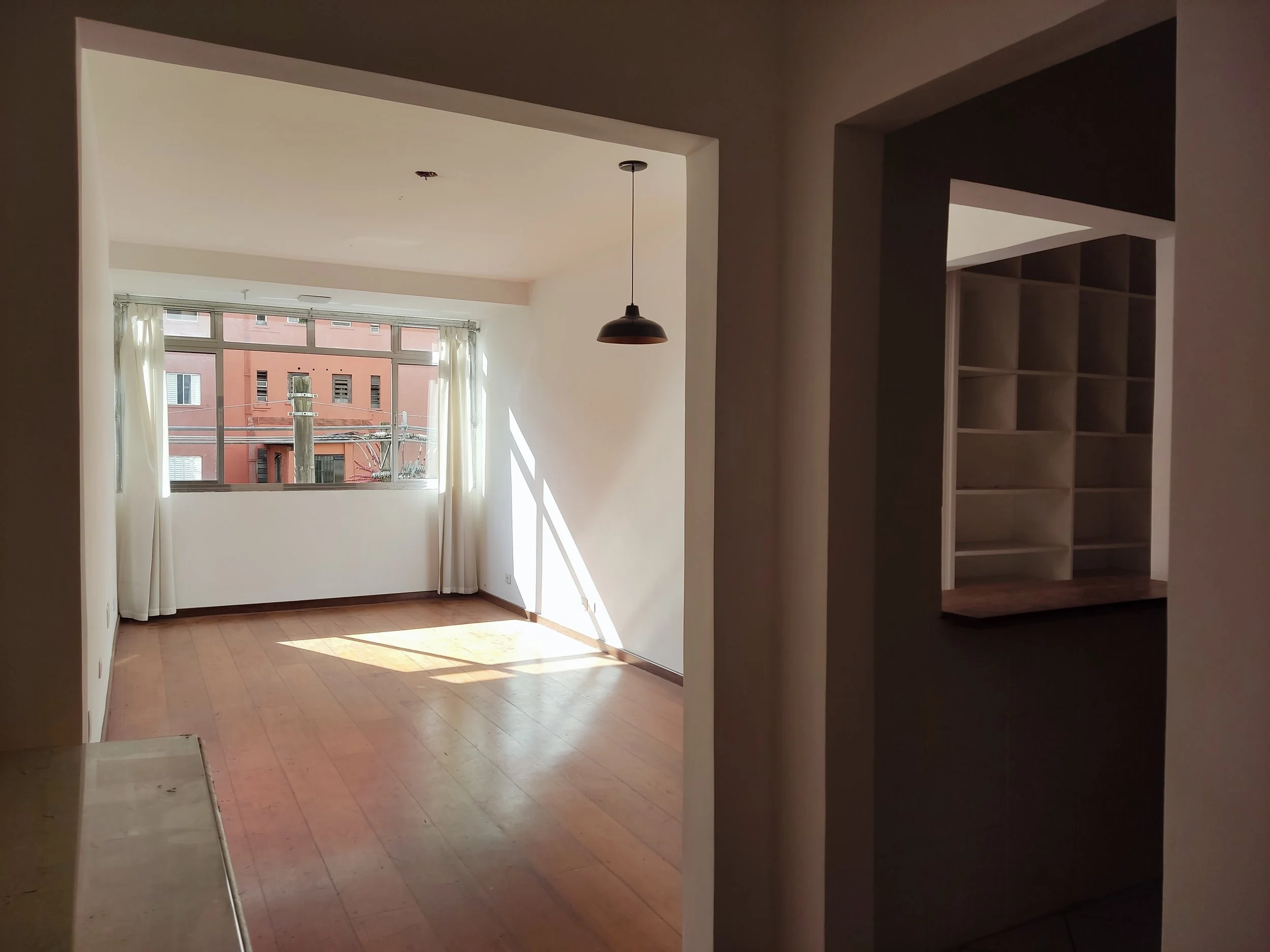 Salas amplas e arejadas com piso de madeira, parede branca, grande janela com cortinas brancas, e iluminação natural confortável.