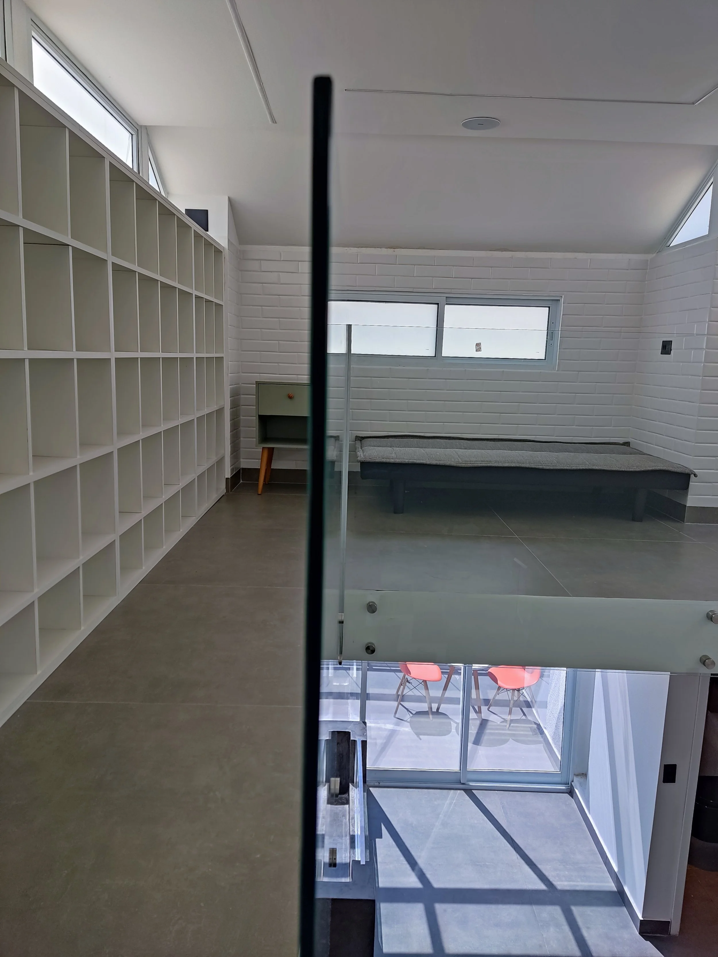 Vista de um ambiente dividido por uma parede de vidro, com uma área superior com cadeiras vermelhas na varanda e uma área interna com uma cama de casal e uma prateleira branca na parede.