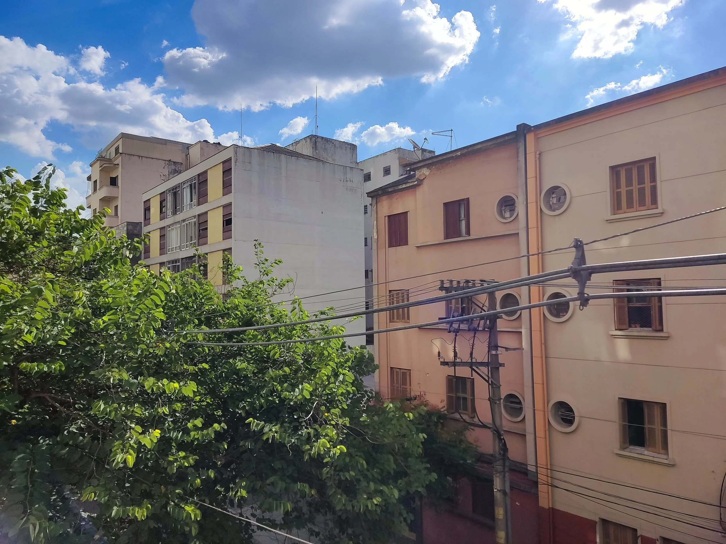 Construções residenciais com árvores verdes na frente, cabos de força e céu com nuvens brancas e céu azul.