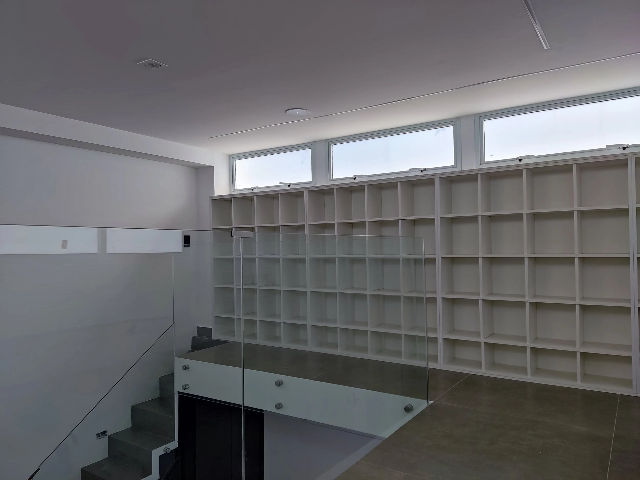 Interior de uma sala moderna com paredes brancas, prateleiras brancas na parede, janelas altas e uma escada com vidro e corrimão metálico.