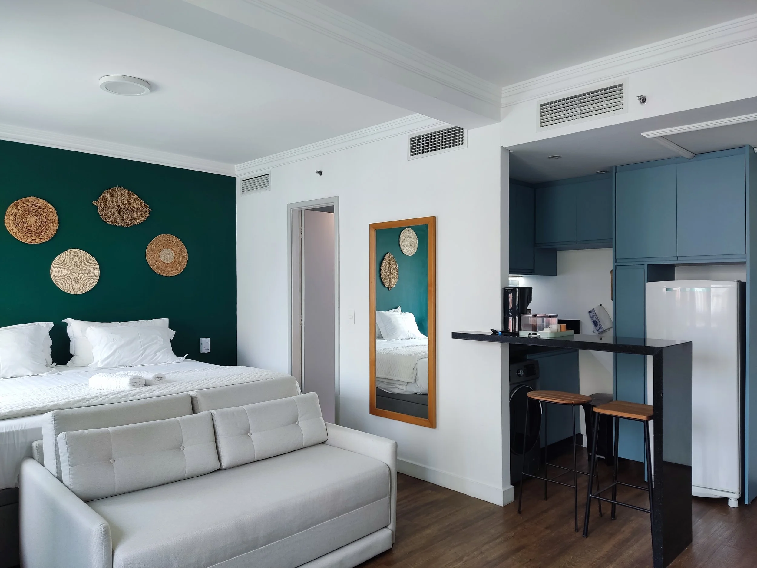 Quarto de hotel com cama com travesseiros e toalhas, parede verde com objetos decorativos, espelho, sofá branco, cozinha com geladeira e banquetas de bar.