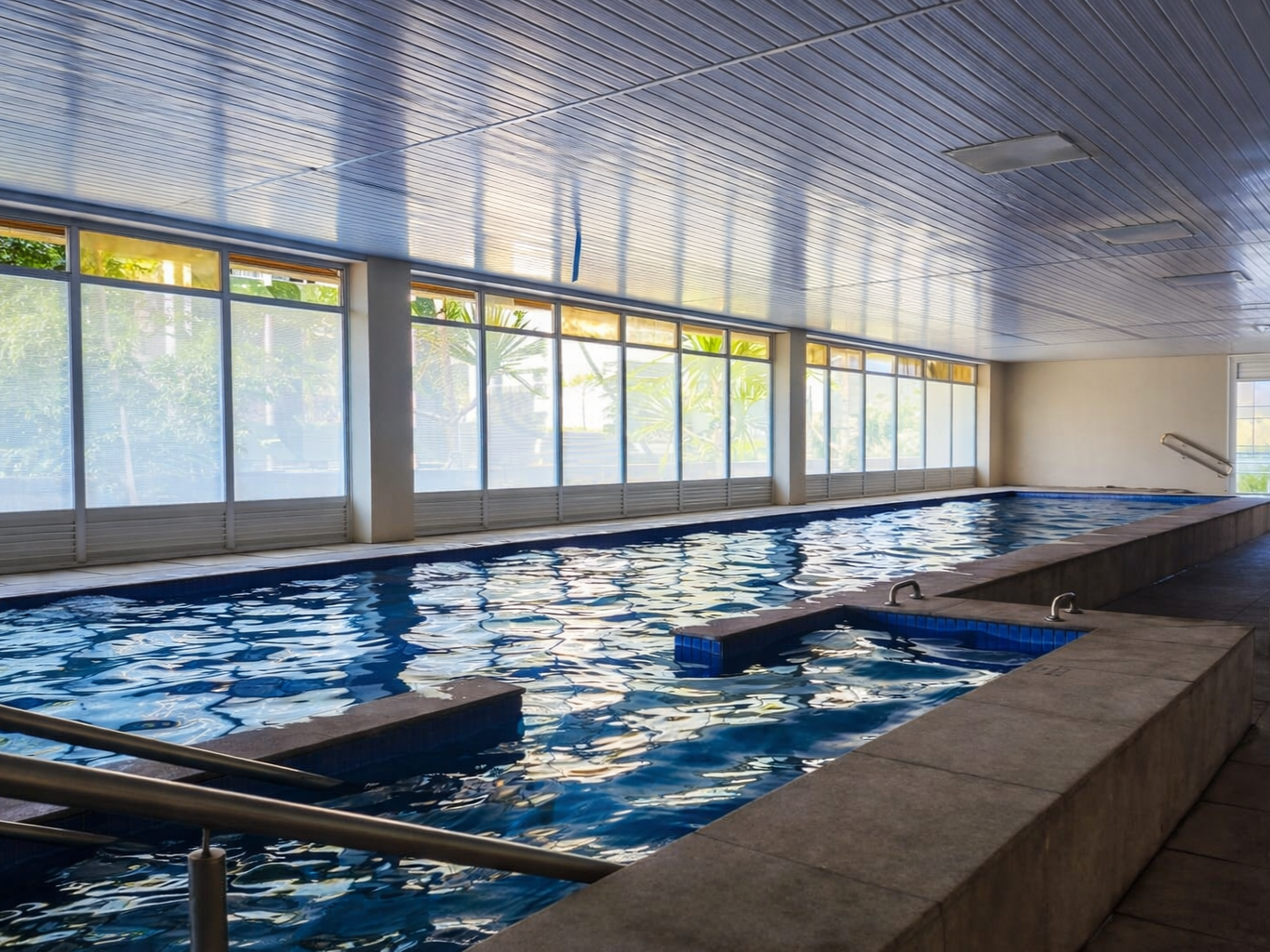 Piscina coberta com janelas do lado, água calma, rampa de acesso e área de descanso ao redor.
