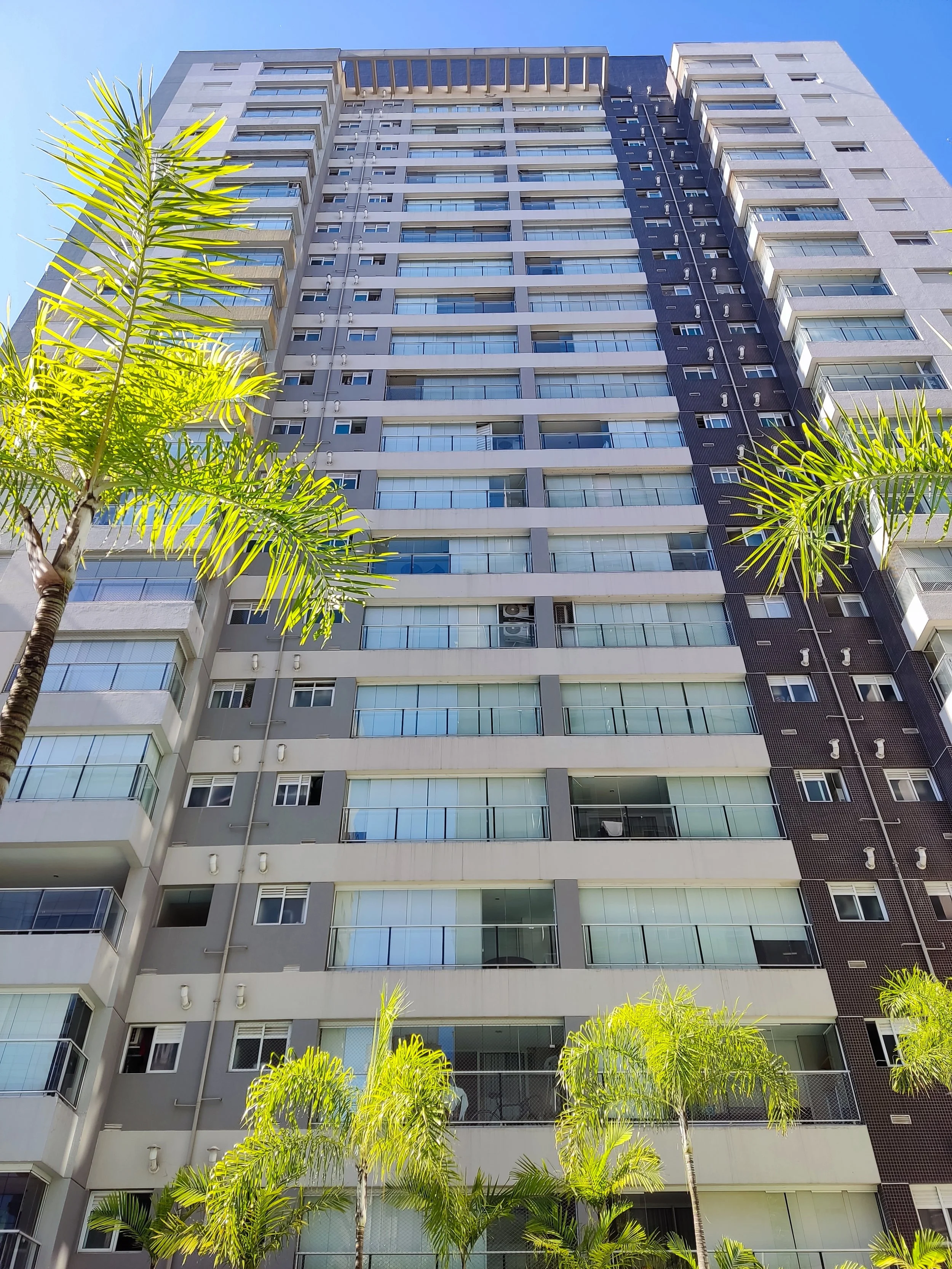 Foto de um prédio de apartamentos alto com várias janelas e varandas, tendo árvores de palmeira na frente, sob um céu azul claro.