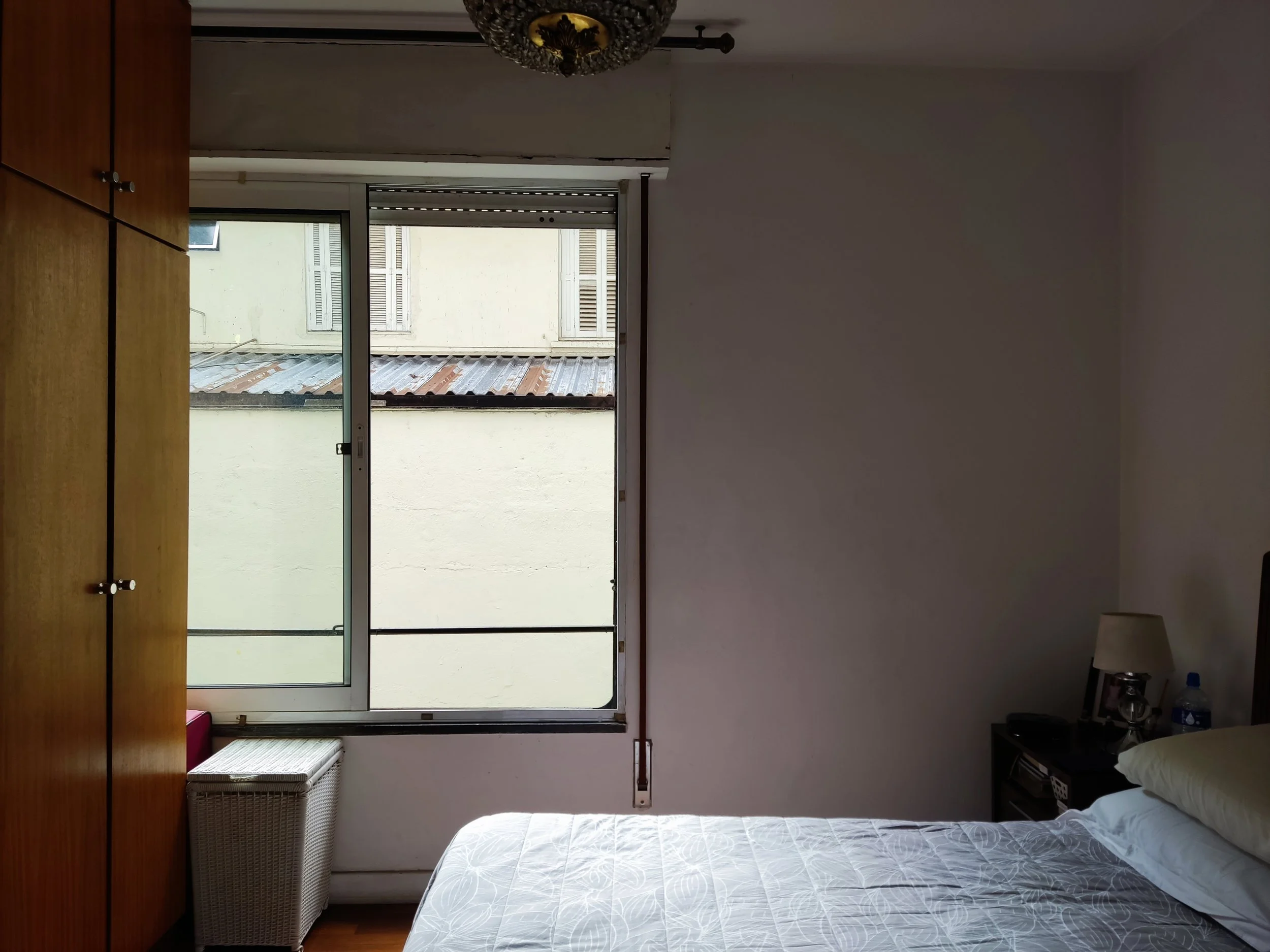 Quarto com uma cama de cama branca, uma cômoda de madeira escura, uma televisão e um criado-mudo com luminária, além de uma janela grande com vista para um muro e prédios ao fundo.