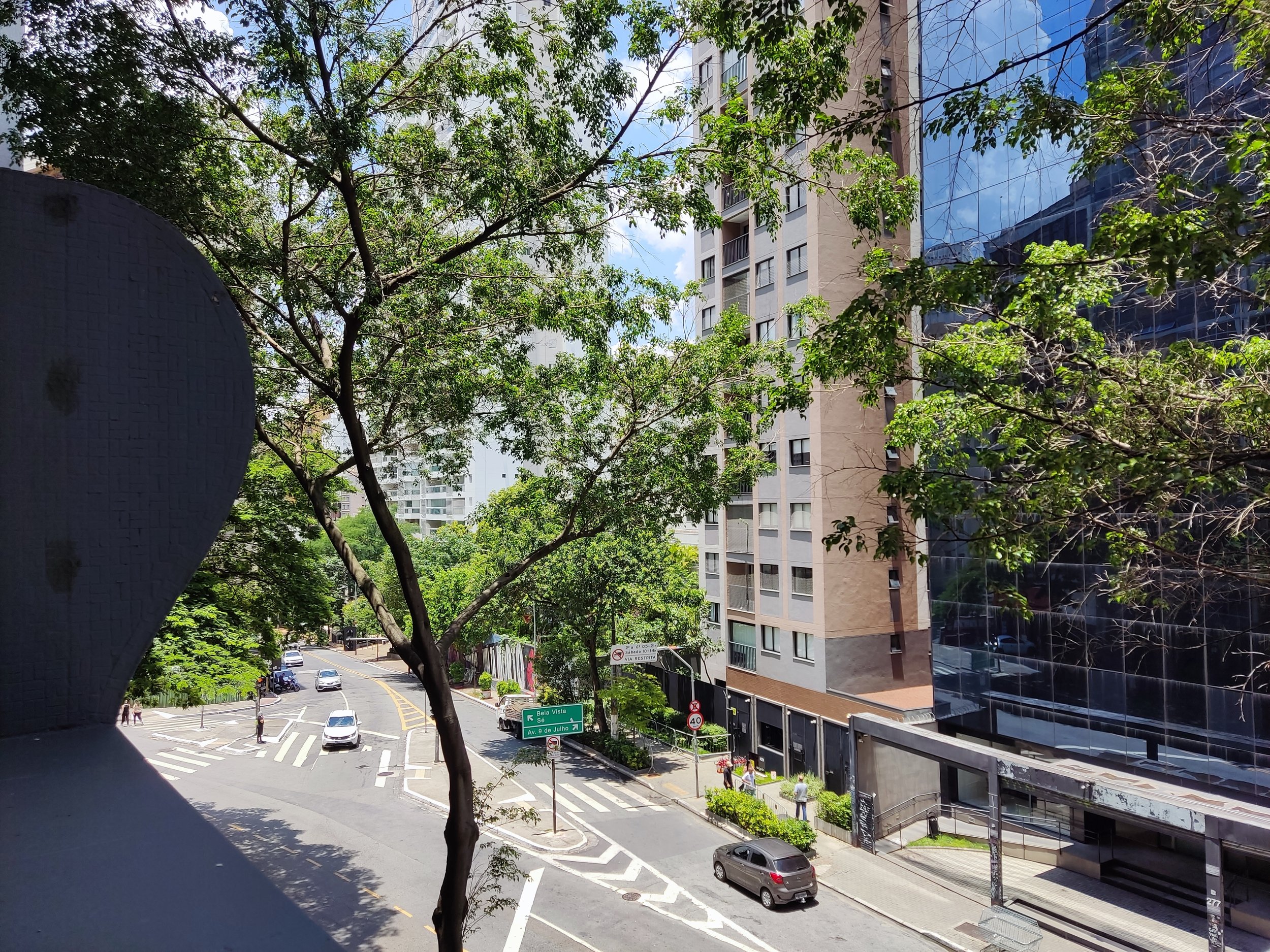 Vista de uma rua urbana com árvores altas, carros, pessoas caminhando e edifícios modernos de vidro e concreto, sob céu azul com algumas nuvens.