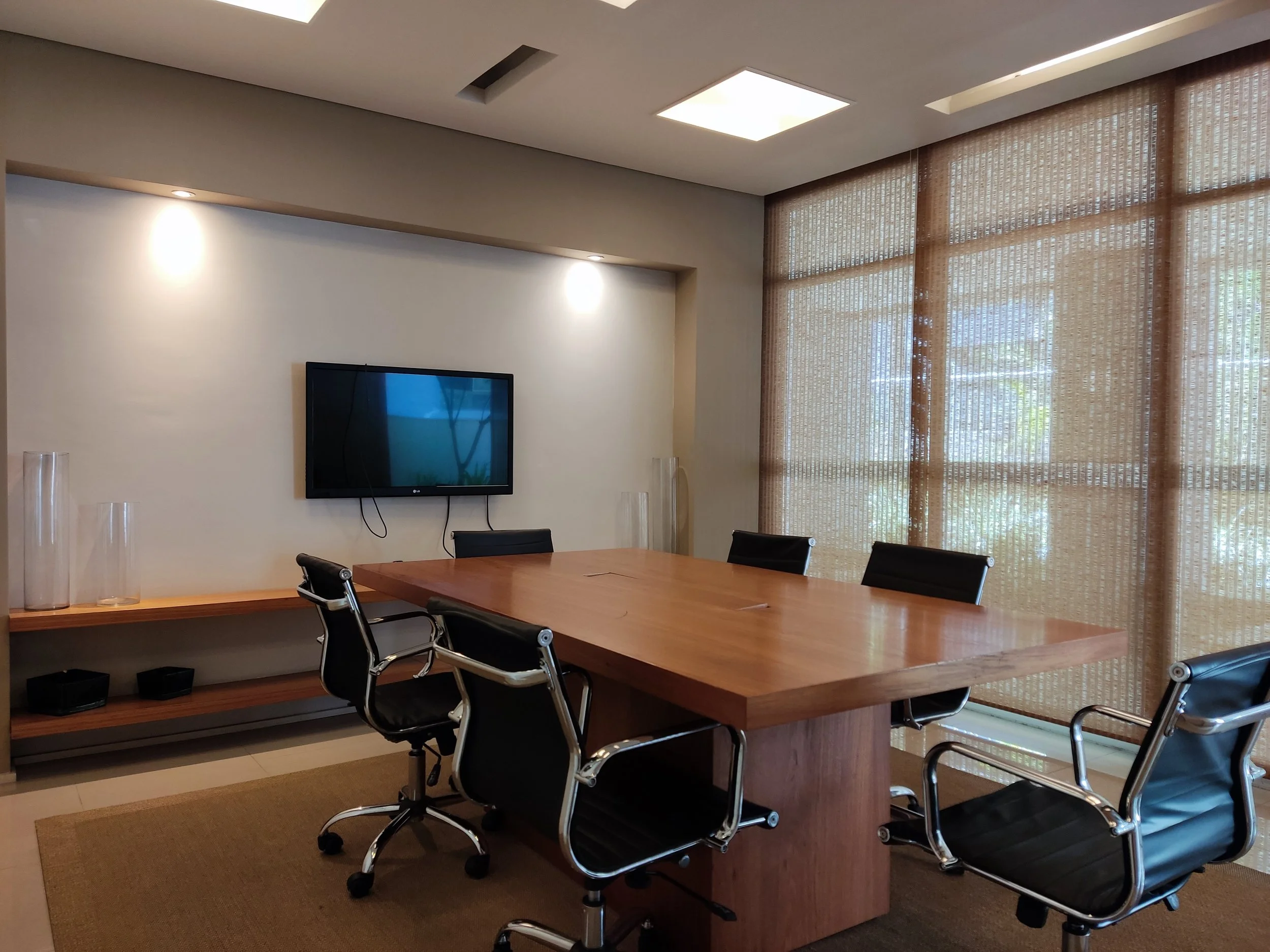 Sala de reunião com mesa de madeira e cinco cadeiras pretas com detalhes em metal, televisão na parede e janela grande com cortinas de bambu.