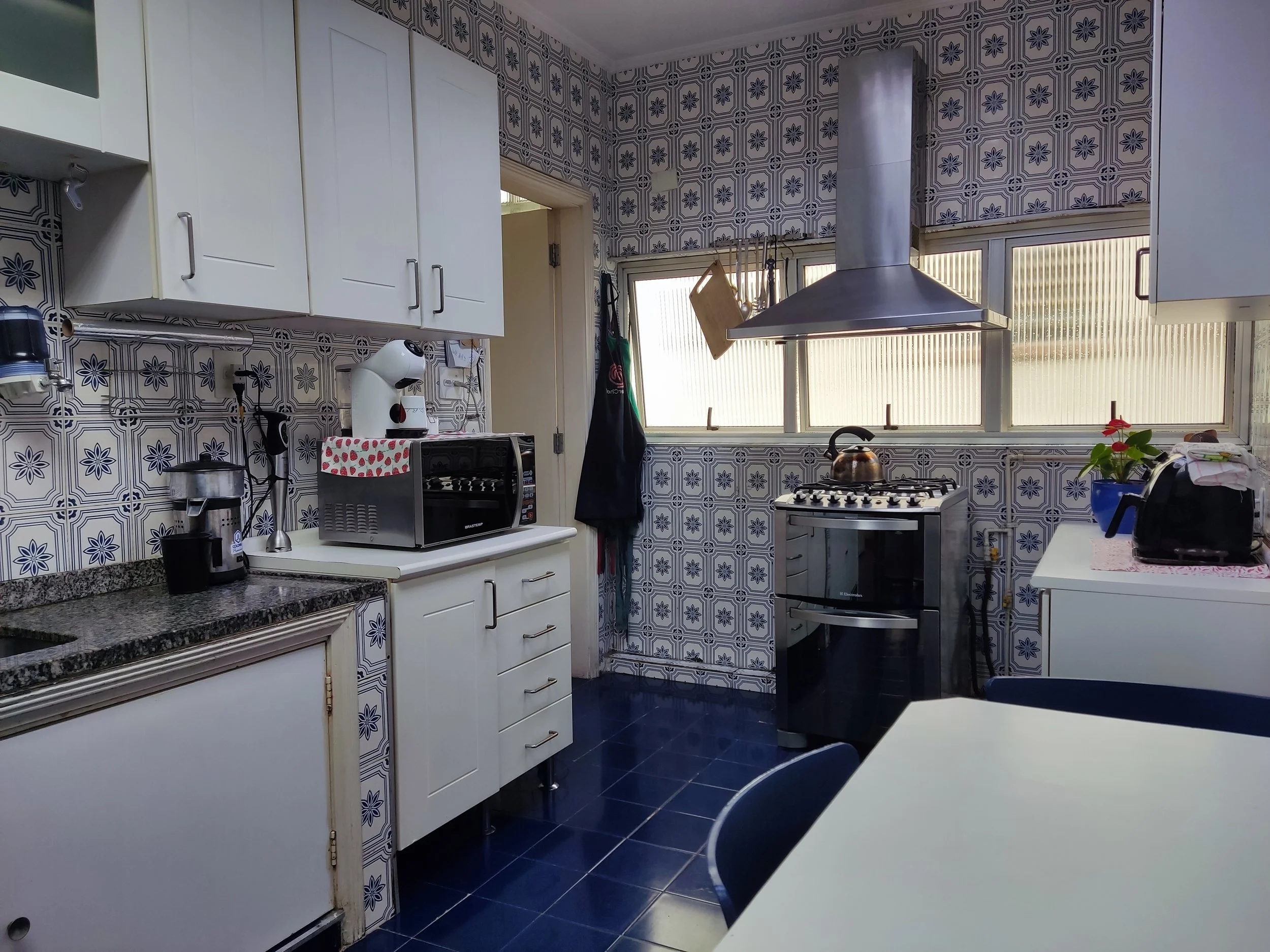 Cozinha com móveis brancos, parede de azulejos decorativos, fogão, forno, utensílios de cozinha, pia, uma planta em vaso, janela com persianas translúcidas, e uma mesa com cadeiras.