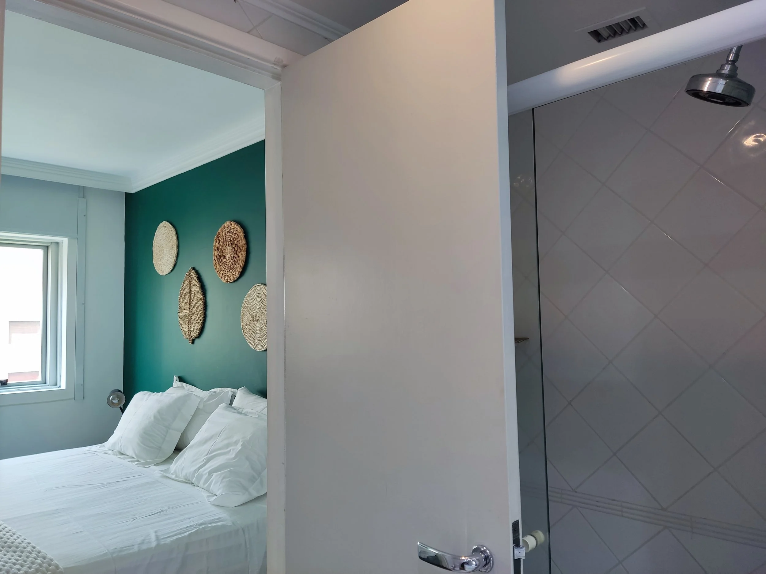 Quarto de cama com parede azul escura decorada com cinco cestas decorativas redondas e gavetas brancas. Ao lado, uma porta aberta revela uma parede de banheiro com azulejos brancos em padrão diagonal e um chuveiro de cabeça fixa.