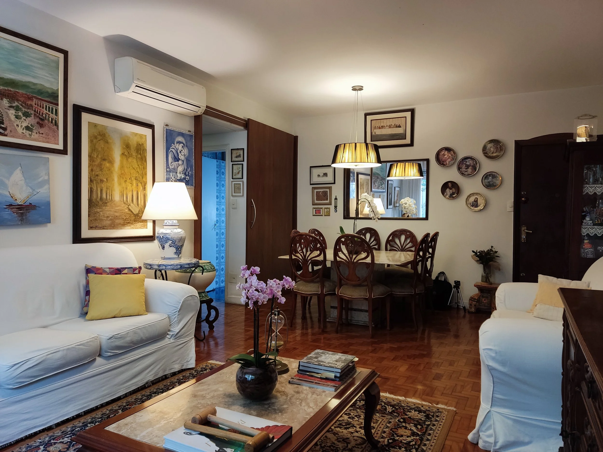 Salão de estar com sofá branco, mesa de centro com livros e vaso de flores, paredes decoradas com quadros e pratos decorativos, mesa de jantar com cadeiras de madeira e luminárias pendentes, parede com espelho e dekoras adicionais.