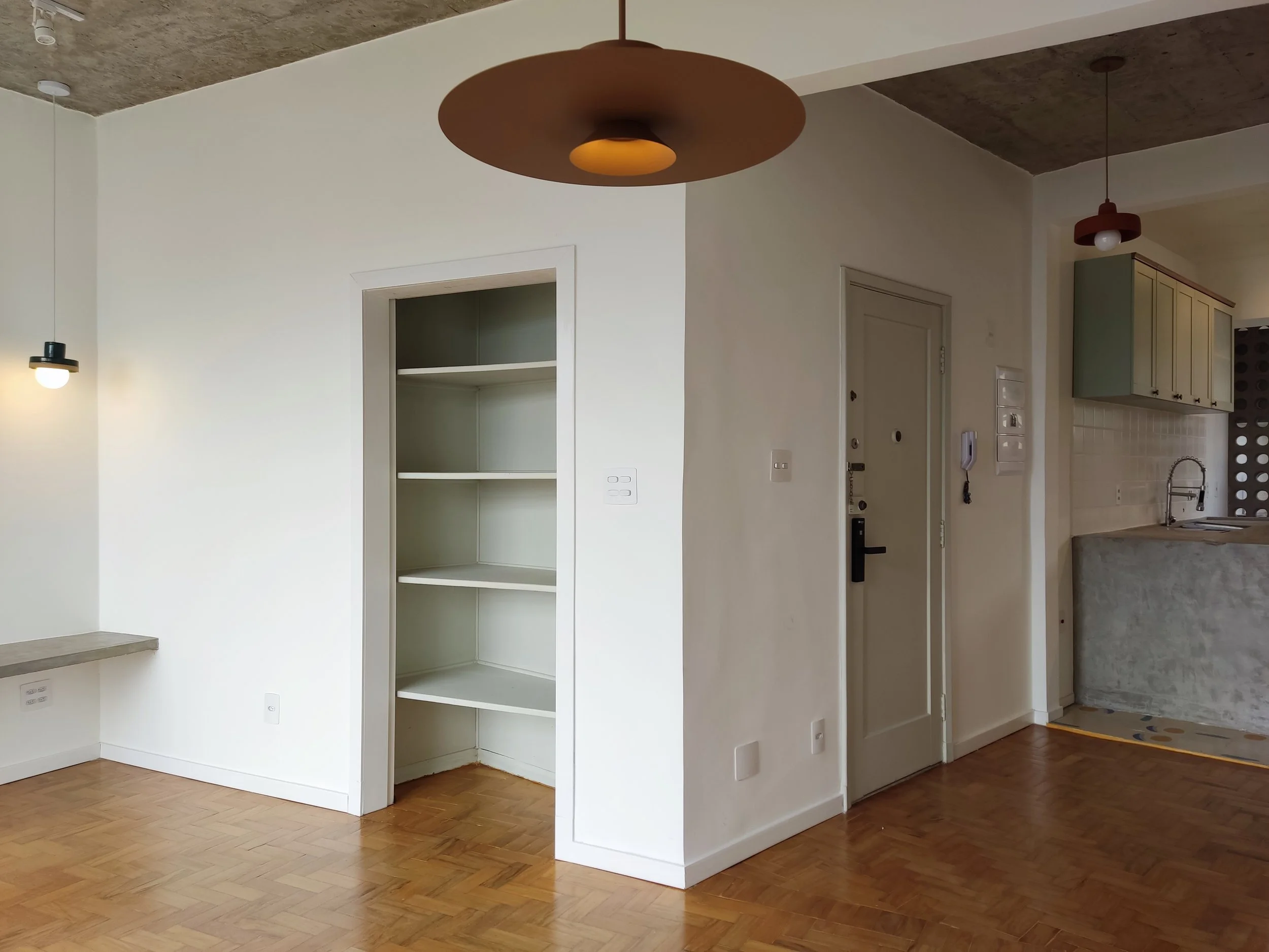 Interior de uma sala com piso de madeira, paredes brancas e iluminação moderna, incluindo uma luminária de centro e uma luminária pendente ao lado da parede.