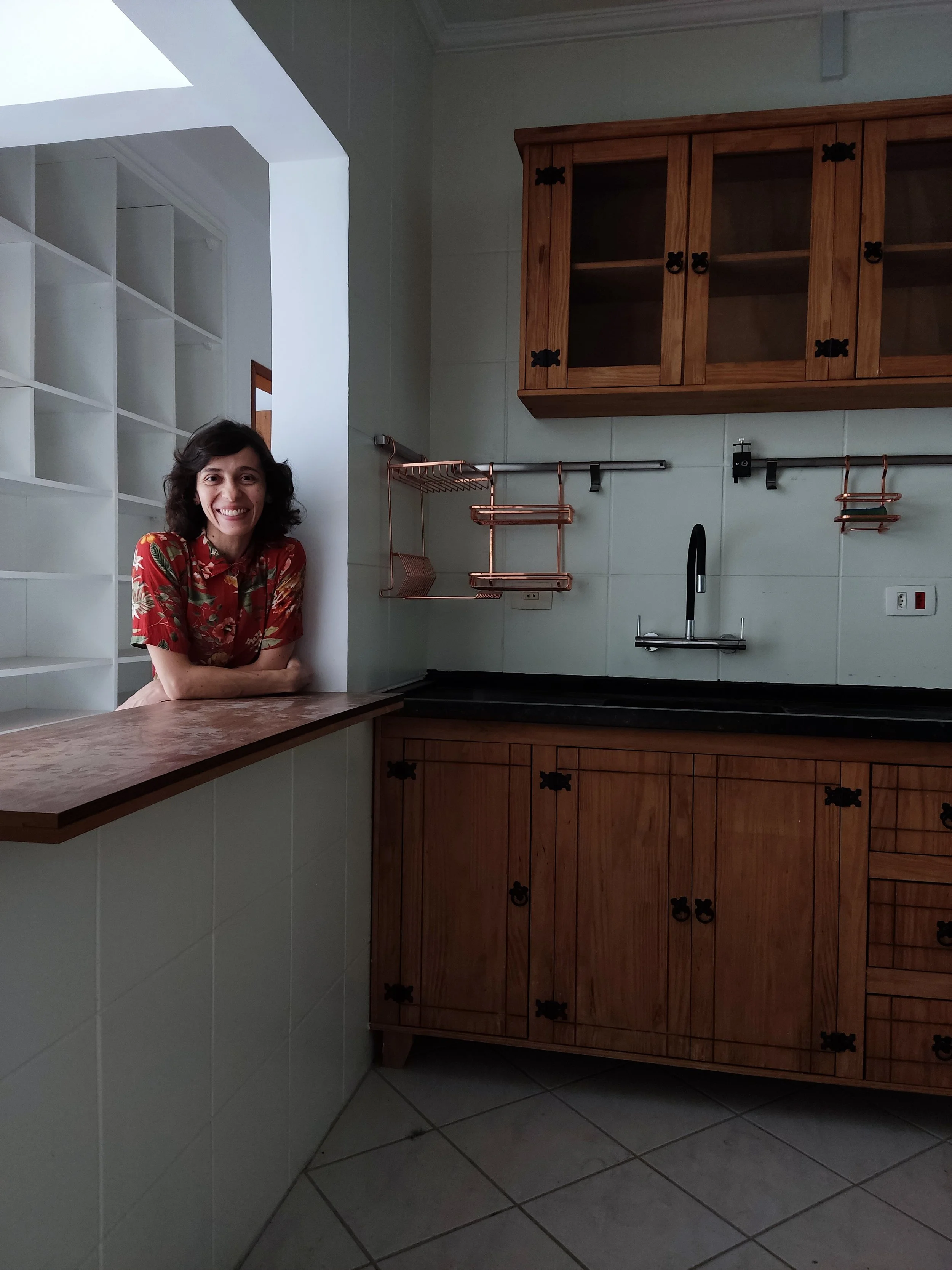 Mulher sorridente com roupa vermelha de estampa floral, apoiada no balcão de uma cozinha com armários de madeira e utensílios de cobre na parede.