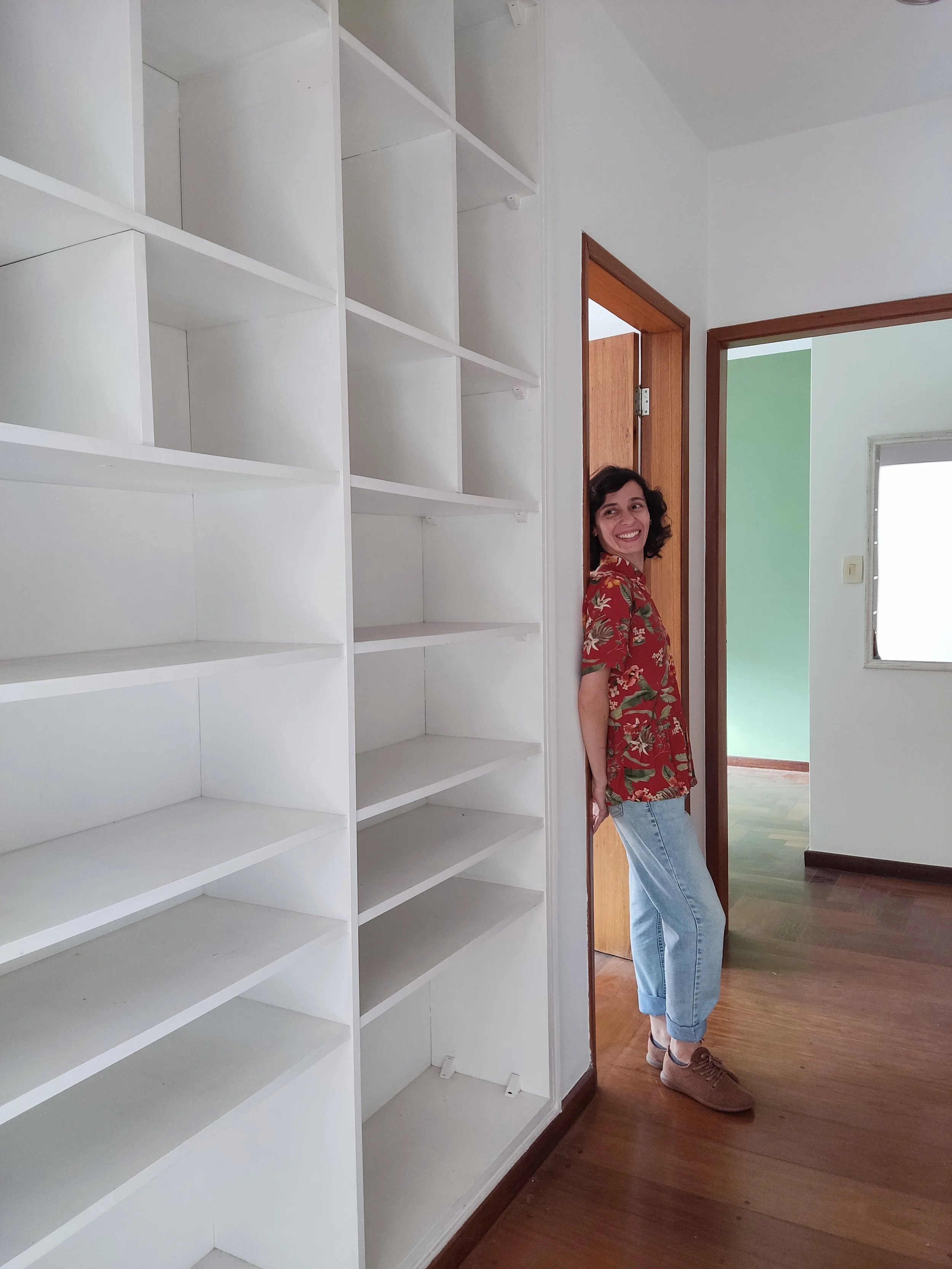 Mulher sorridente de camiseta floral vermelha e calça jeans apoiada na porta de um armário de estantes brancas, em uma casa com piso de madeira e paredes brancas.