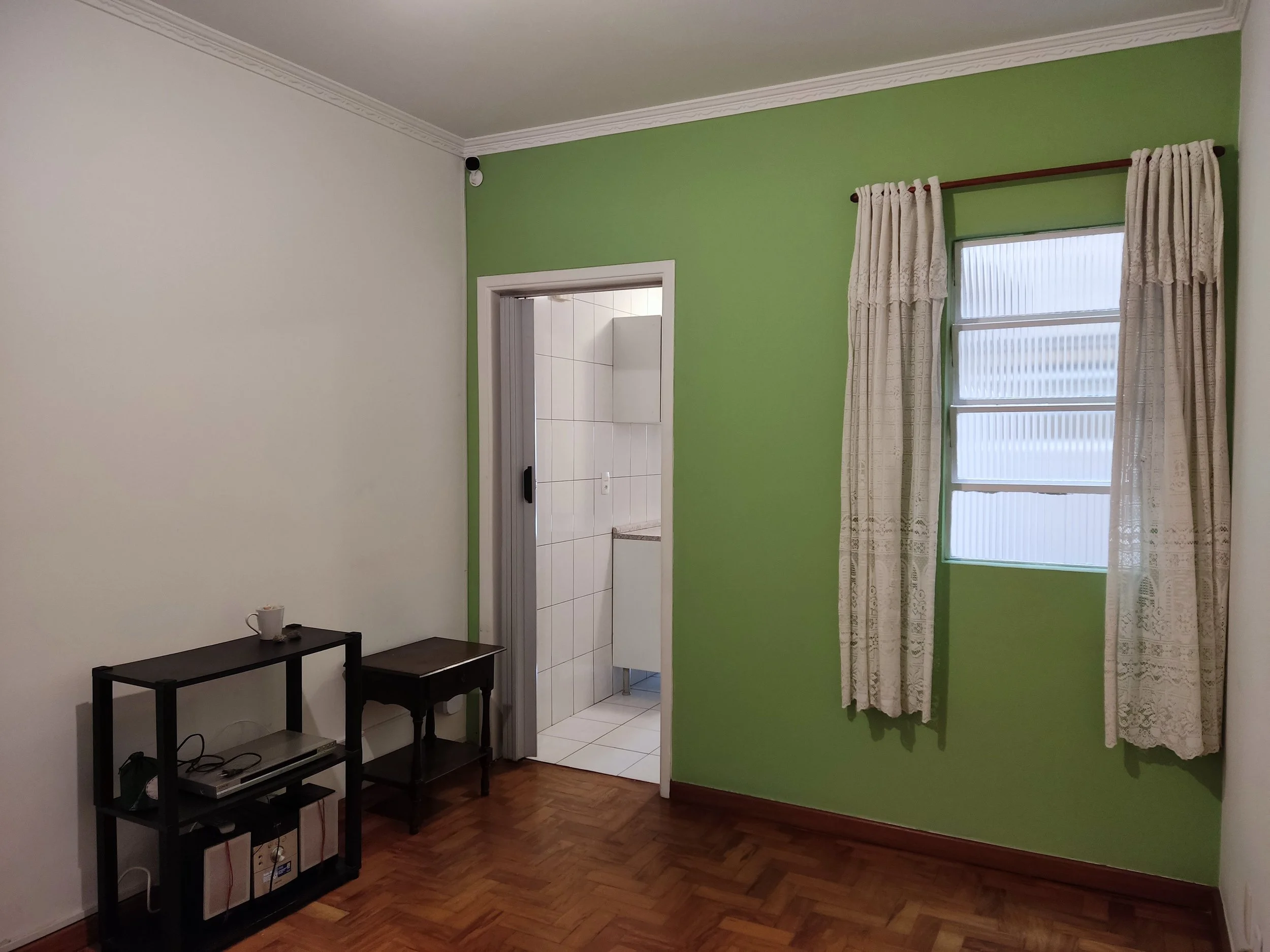 Quarto com parede verde, janela com cortinas brancas, porta que leva ao banheiro e móvel de sala com eletrônicos e livros