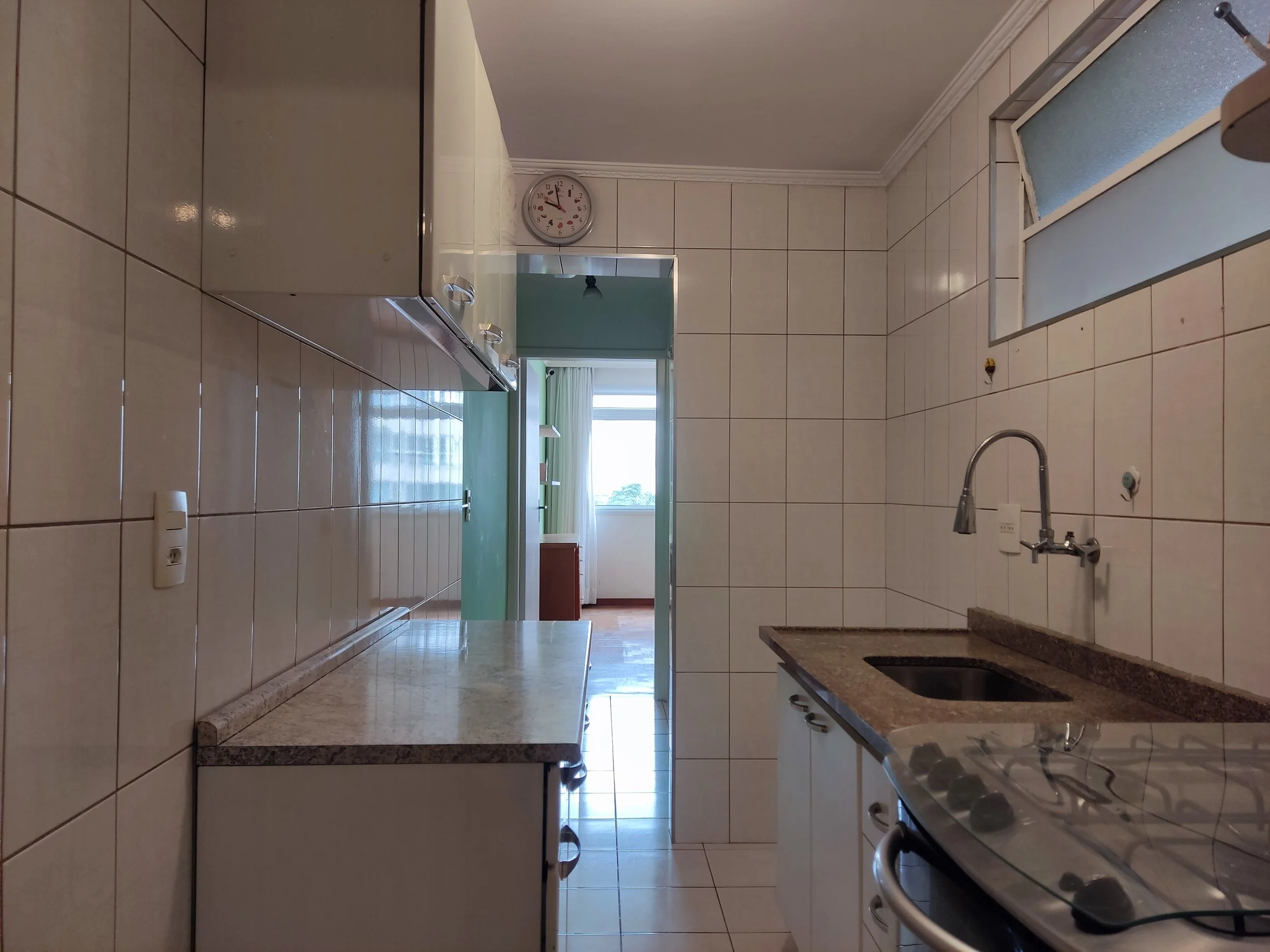 Cozinha com paredes de azulejos brancos, pia de granito com fogão de inox ao lado, armários brancos, relógio na parede e uma janela com vidro fosco. Ao fundo, uma sala com janela grande e cortinas brancas.