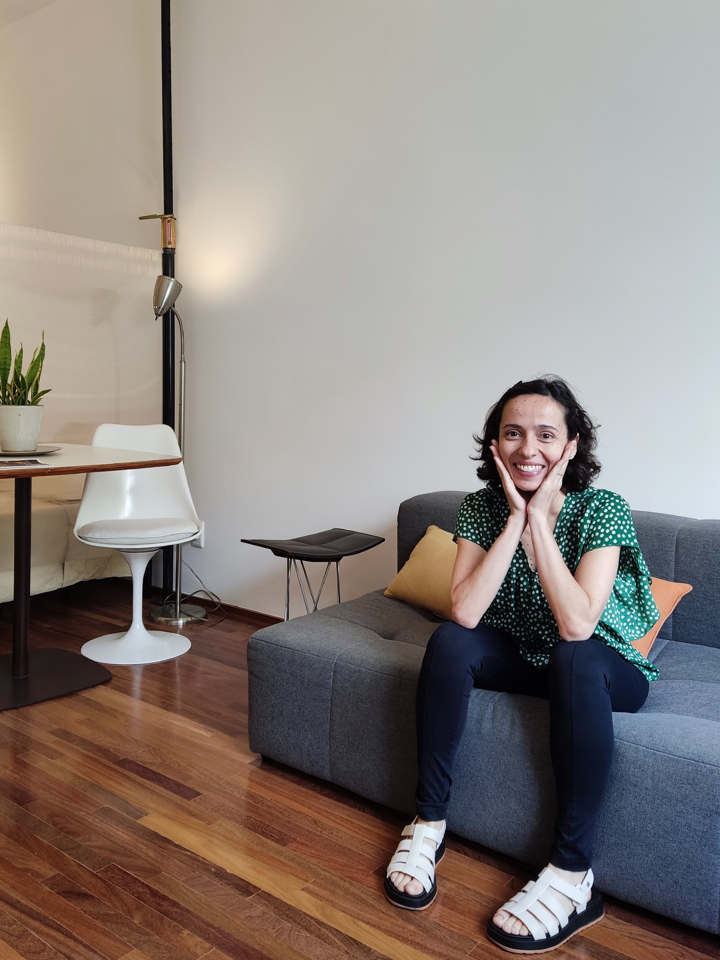 Mulher sorridente sentada em um sofá na sala, com cabelo cacheado, usando calça escura, camiseta verde com branco, sandálias brancas e observed no fundo uma mesa com plantas e uma luminária.