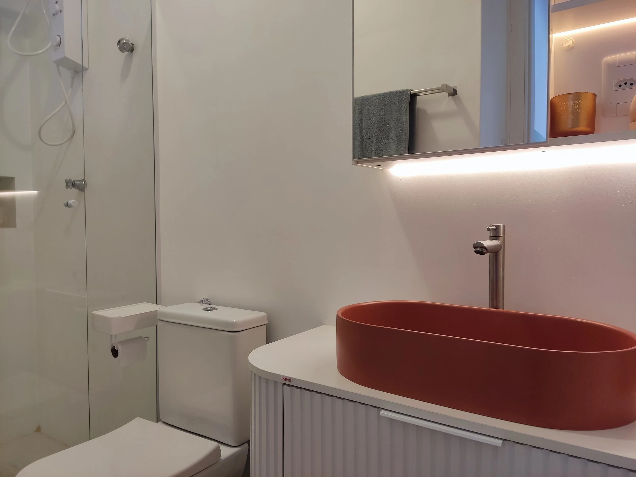 Banheiro moderno com vaso branco, pia com cuba grande de cor terracota, espelho com iluminação embutida, toalheiro com toalha cinza, vidro de banho e parede branca.