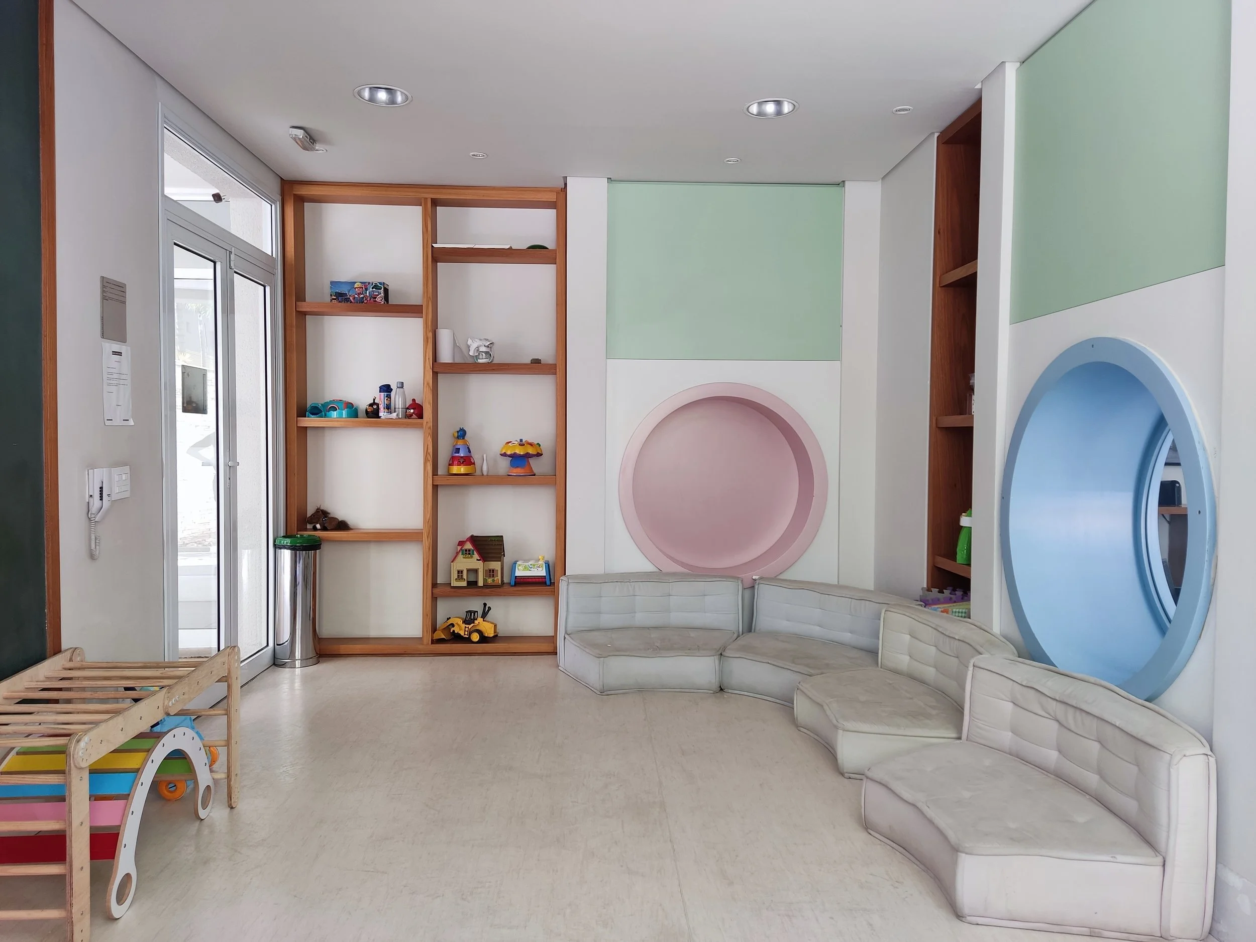 Sala de espera de uma clínica ou consultório infantil com sofá em forma de U, prateleiras de madeira com brinquedos, paredes decoradas com cores pastel azul, rosa e verde, e uma porta de vidro.