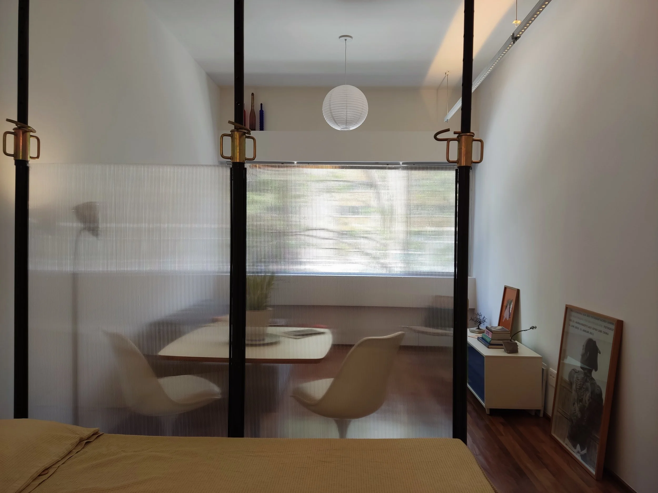 Divisão de espaço com vidro com textura, sala de jantar com mesa e cadeiras ao fundo, e um canto com prateleira e quadros encostados na parede.