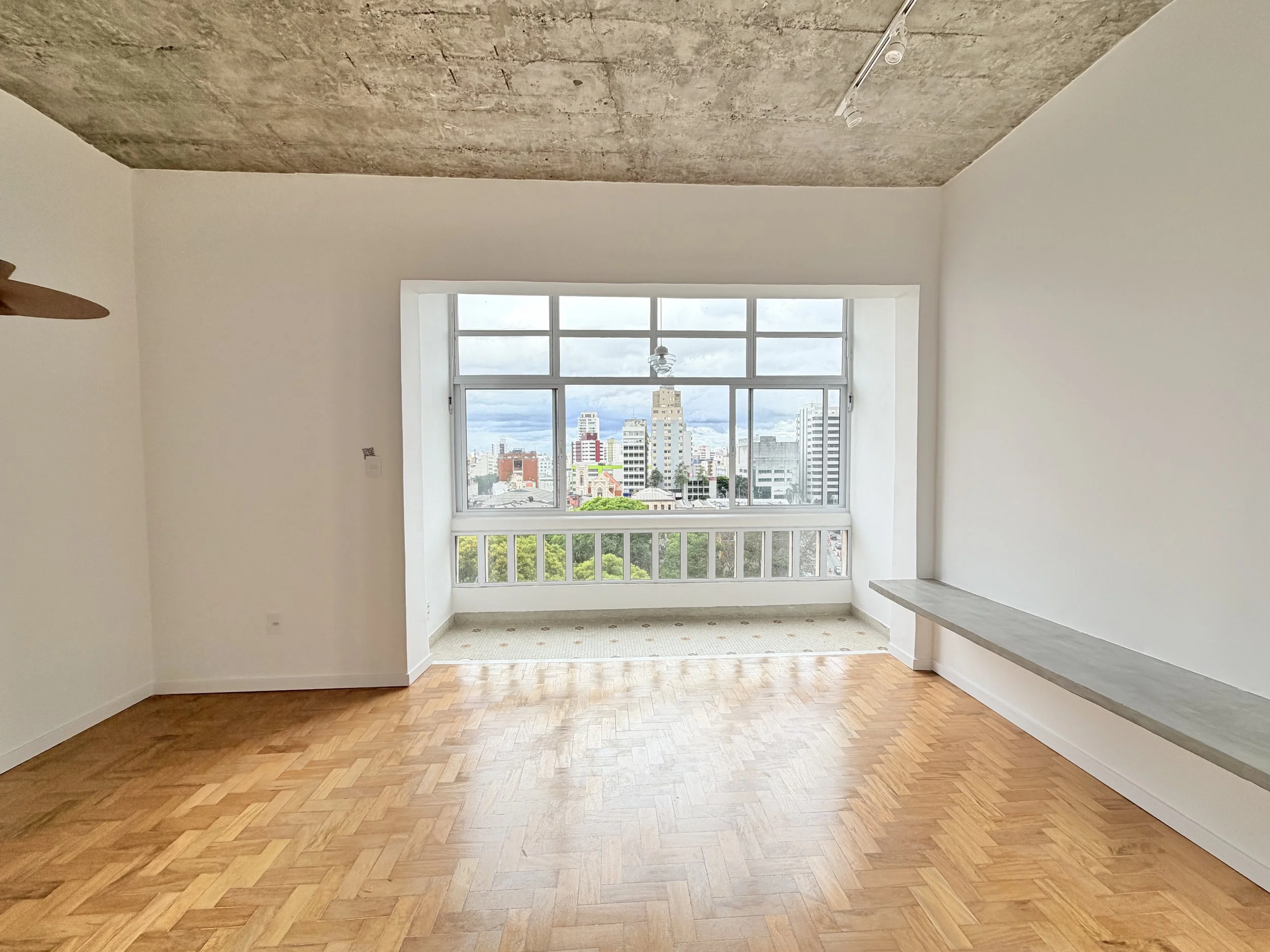 Sala vazia com piso de parquet, paredes brancas, janela grande com vista para uma cidade e teto de concreto exposto.