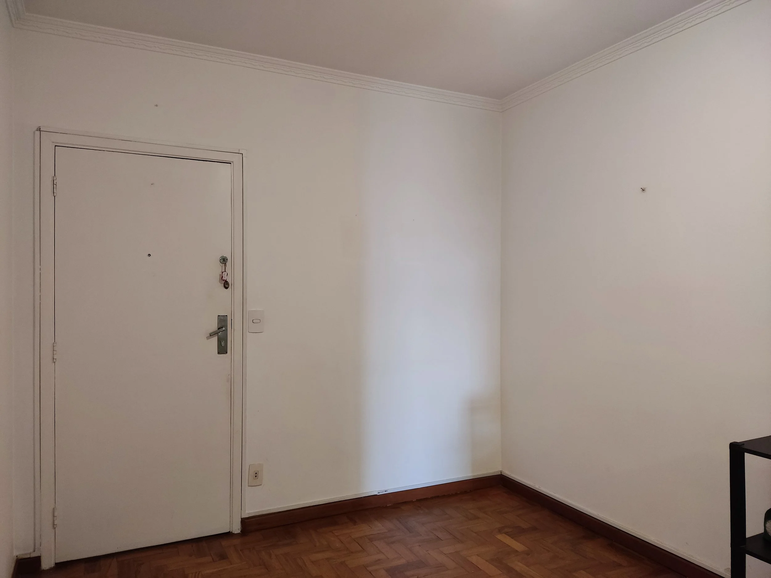 Parede branca com porta de entrada cinza e piso de madeira, espaço vazio com uma pequena mesa preta no canto direito.